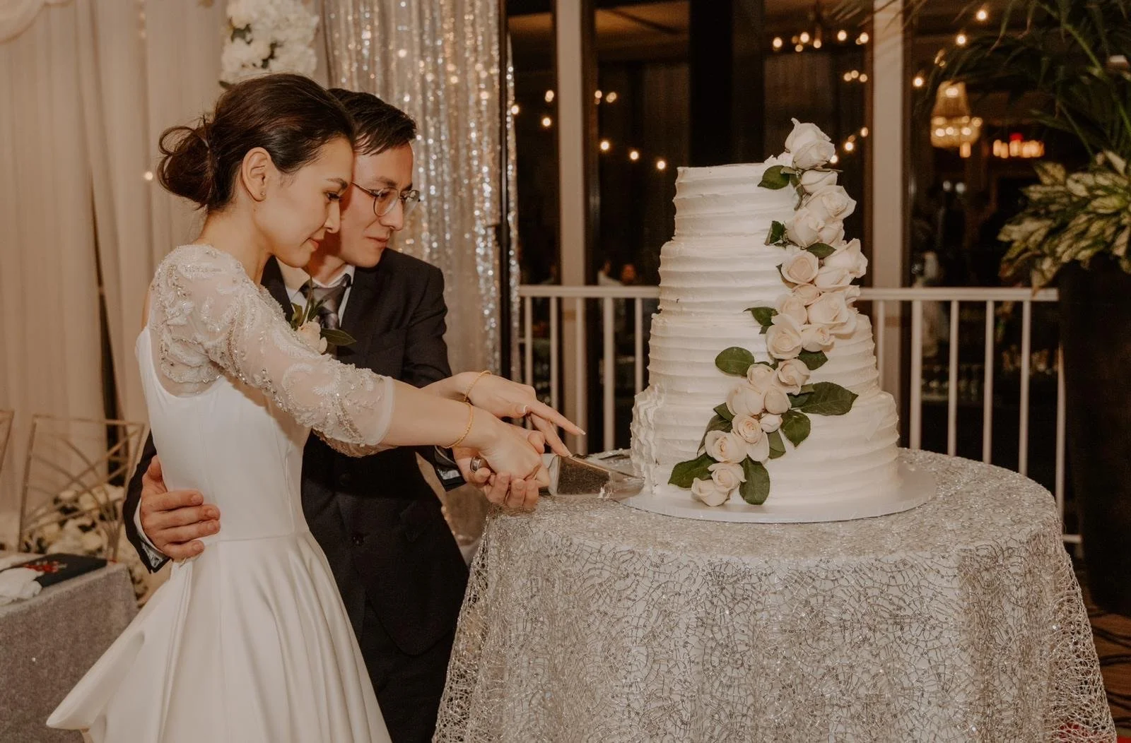 A bride and groom cutting a wedding cake together at their wedding reception, with a multi-tiered white cake decorated with pink roses and green leaves.