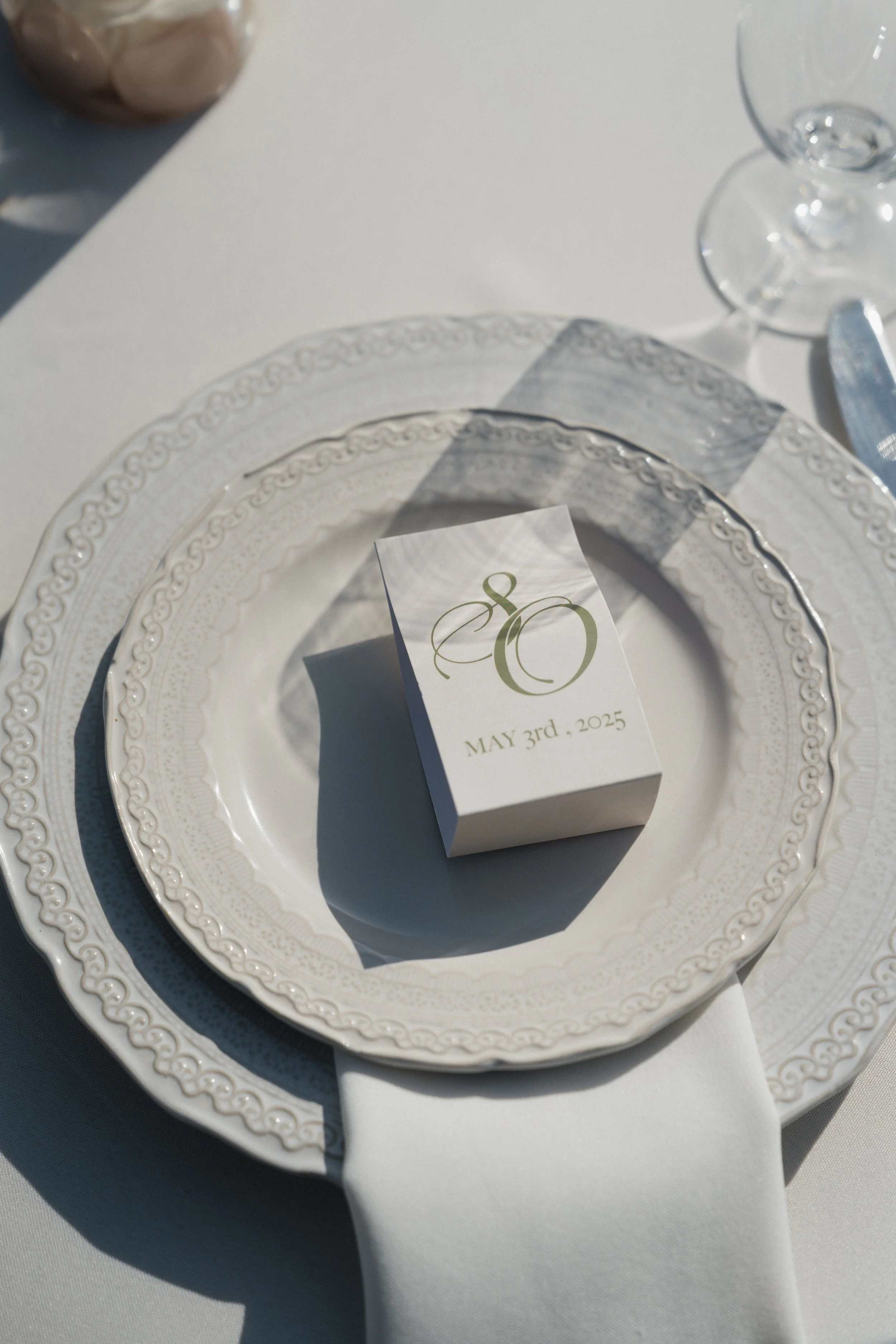Elegant wedding table setting with a white plate, a decorative charger, a folded napkin, and a small box with the date May 3rd, 2025, on a white tablecloth.