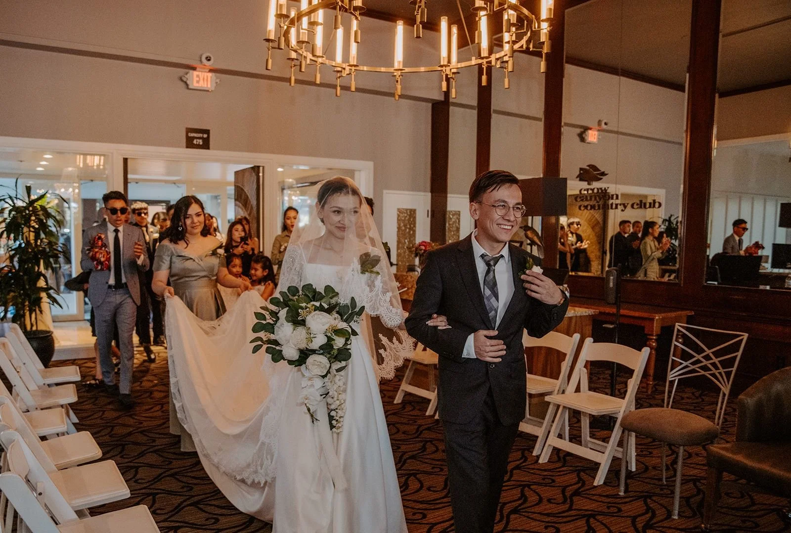 A wedding ceremony with a bride in a white dress and veil holding a bouquet walking down the aisle, accompanied by a groom in a suit. Guests are in the background, some taking photos and smiling, in a warmly lit indoor venue with chairs and a chandel