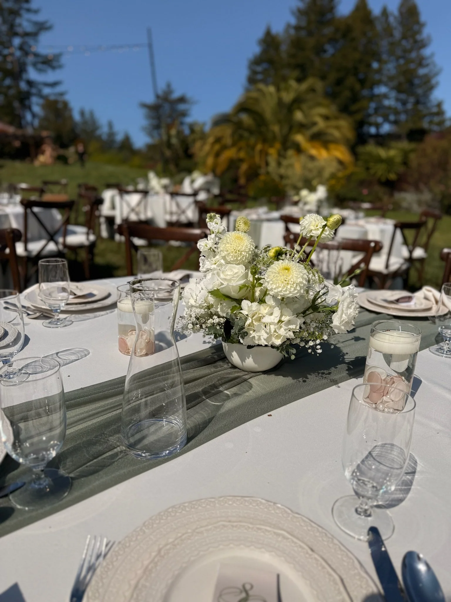 Elegant outdoor table setting with a floral centerpiece, surrounded by white plates, glasses, and cutlery, with trees and a clear sky in the background.