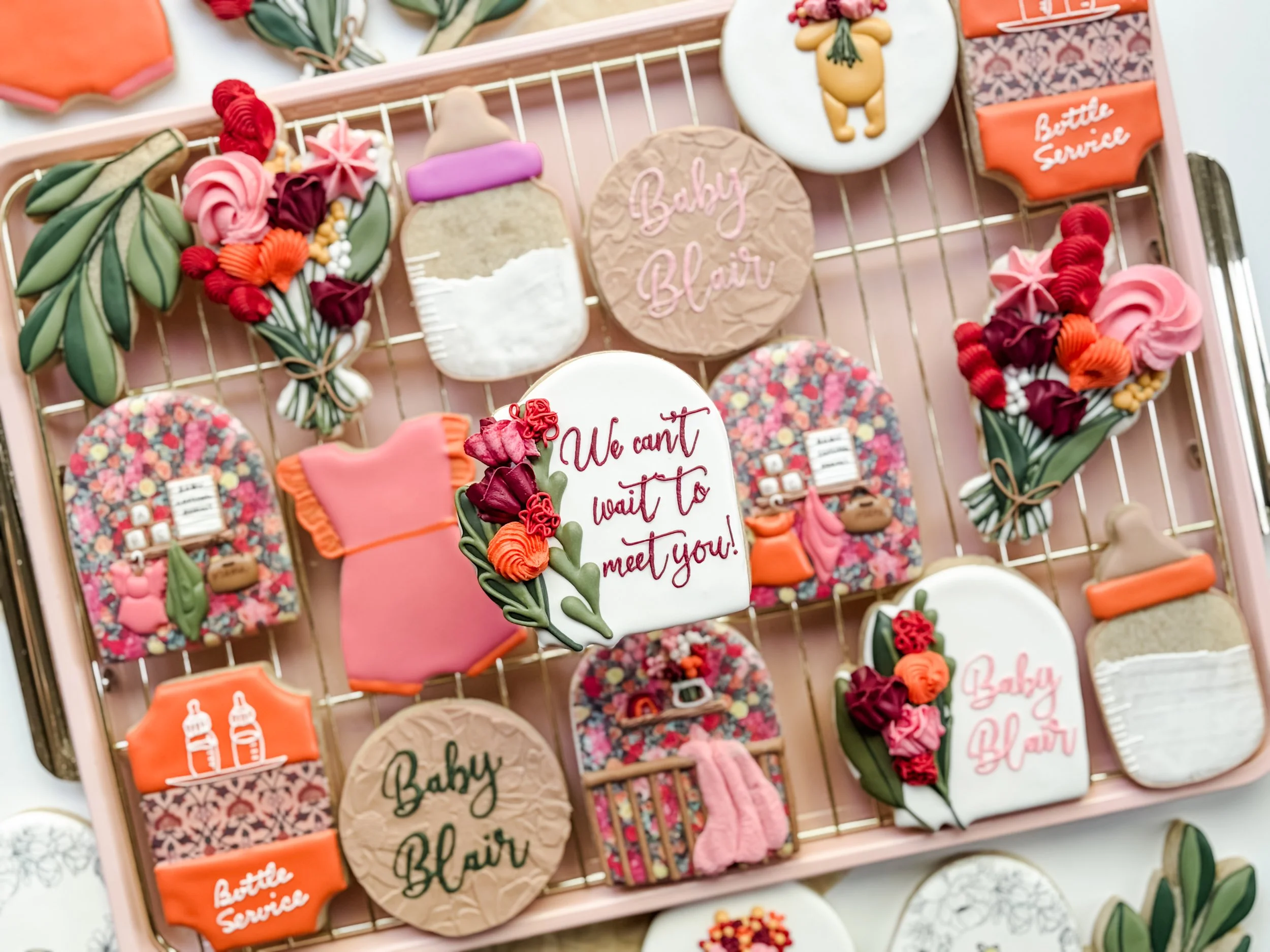 Decorated cookies with various designs and messages, including cookies with flowers, baby bottles, the words 'Baby Blair', and 'We can't wait to meet you!' on pink and beige backgrounds, displayed on a pink tray.