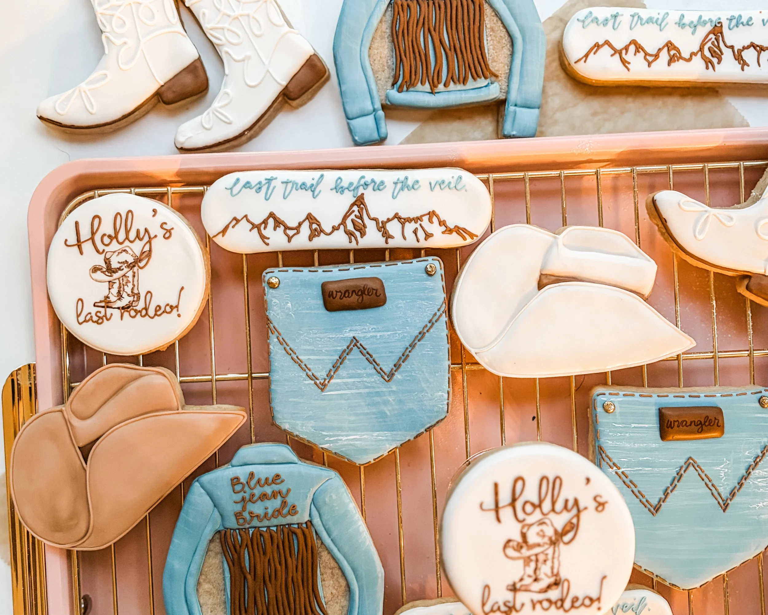 Various intricately decorated cookies in a camping and outdoor theme, including ice skates, a mountain landscape with a trail, a message reading 'Last trail before the veil,' Wrangler jean pockets arranged on a pink tray.