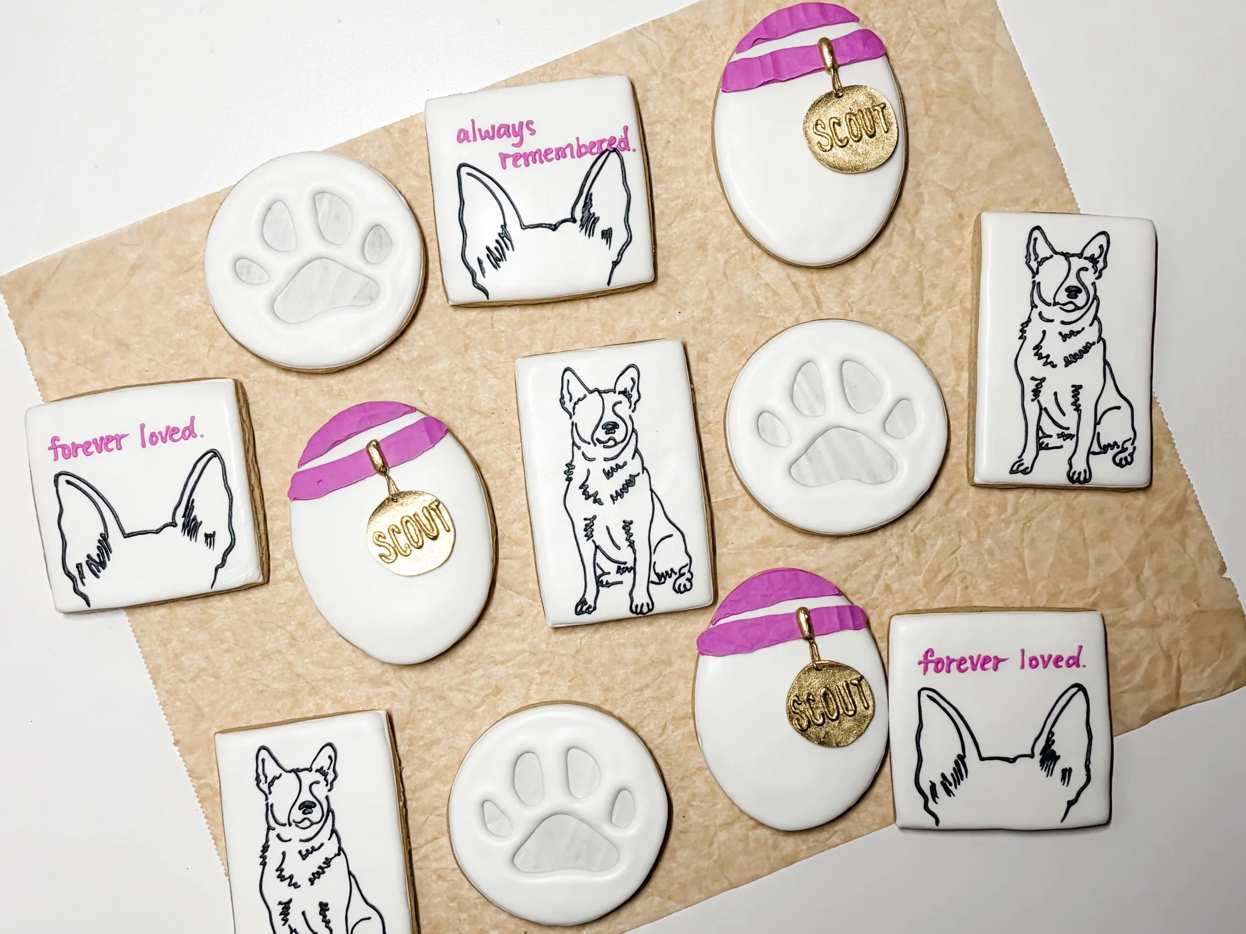 Decorated cookies with dog images, paw prints, and phrases like "always remembered," "forever loved," and "SCOUT," placed on a tan parchment paper. Some cookies have pink and purple accents.