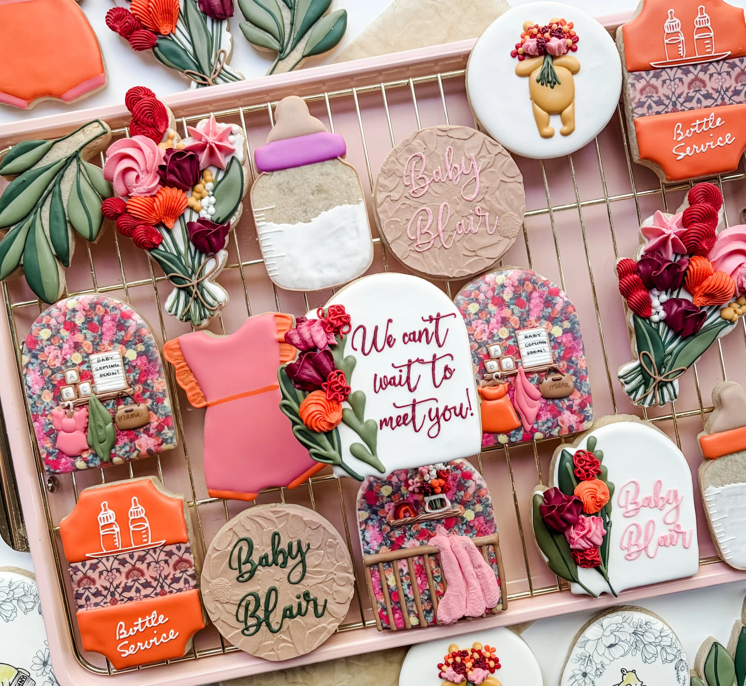 Assorted decorated cookies with baby and name themes, including baby bottles, flowers, and the words 'Baby Blair' and 'We can't wait to meet you' on pink, white, and patterned backgrounds, arranged on a pink display rack.