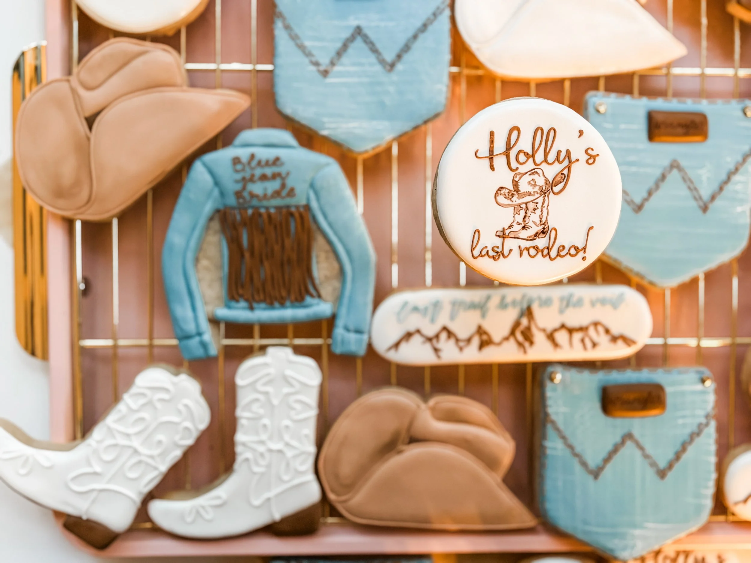 Decorative cookies shaped like cowboy hats, boots, and lassos, with some cookies featuring words like 'Holly's last rodeo!' and images of a cowboy hat and a mountain range.