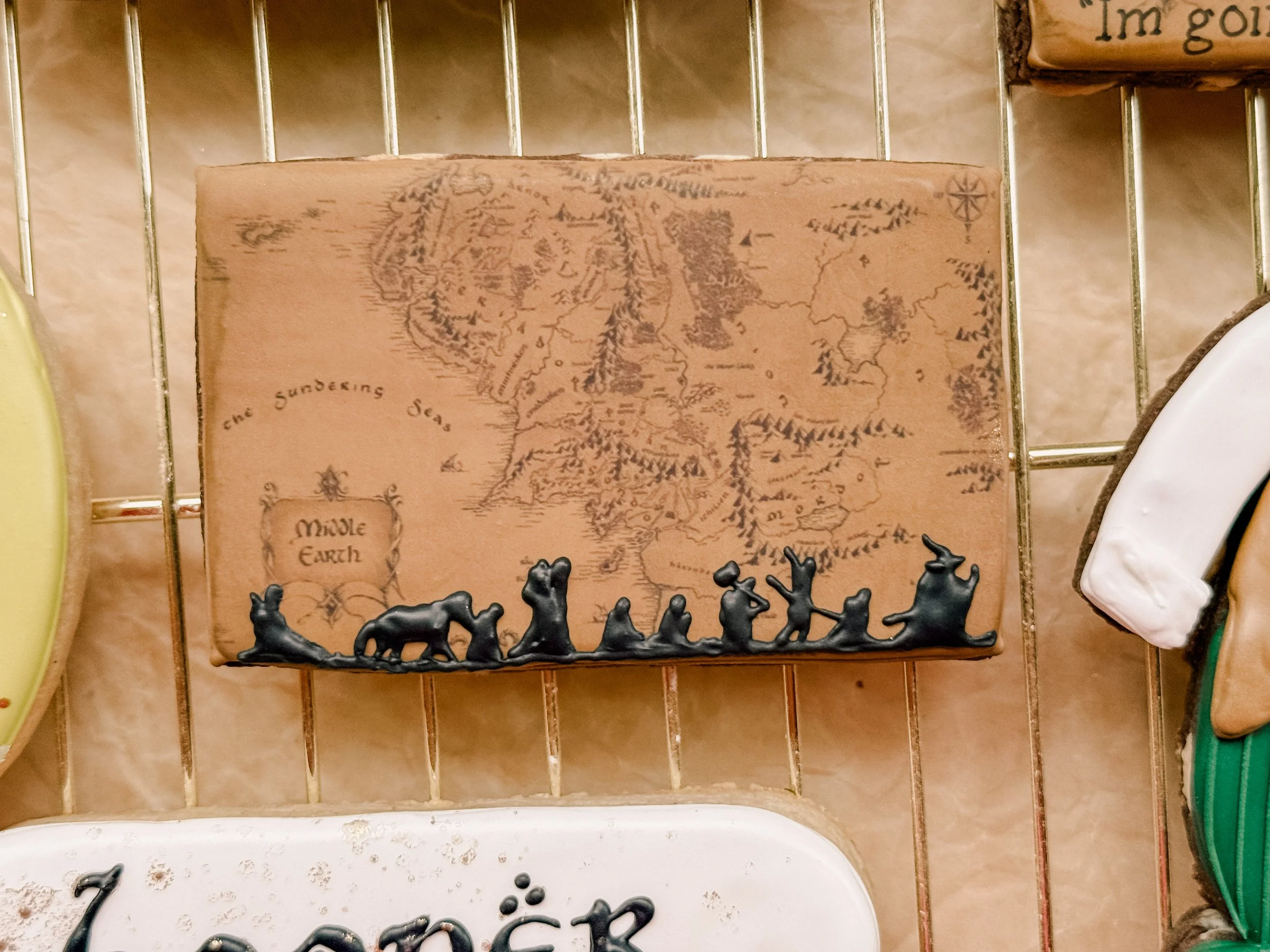 A vintage-style map of Middle Earth attached to a pipe or shelf backing, with black decorative dragon-shaped objects in front of a white space. Part of a sign with partial text is visible in the top right corner.