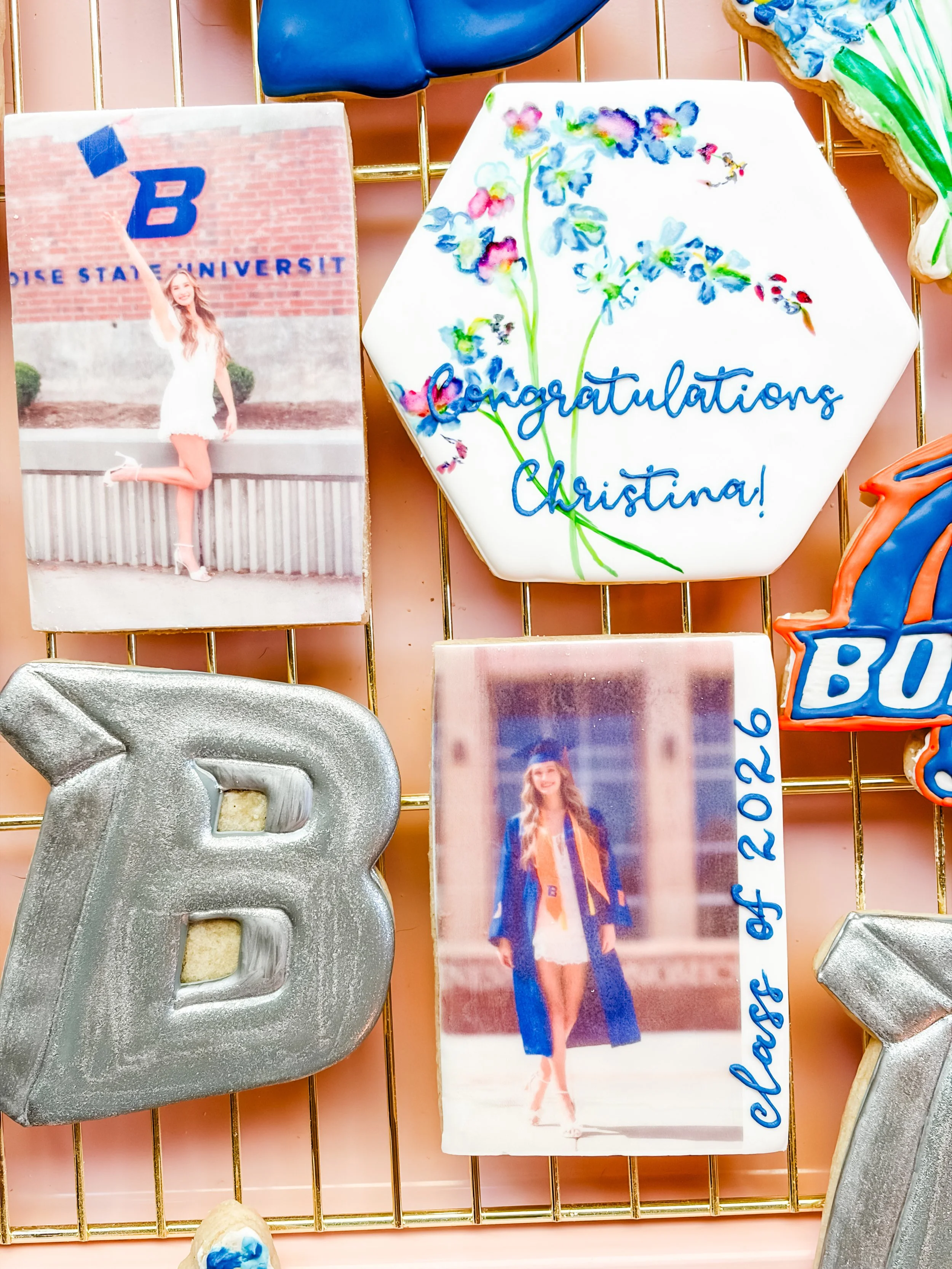 A decorated cookie with white icing, colorful painted flowers, and the message 'Congratulations Christina!' written in blue. There are also graduation photos and a glittery '2026' decoration and a silver 'B' letter on display.