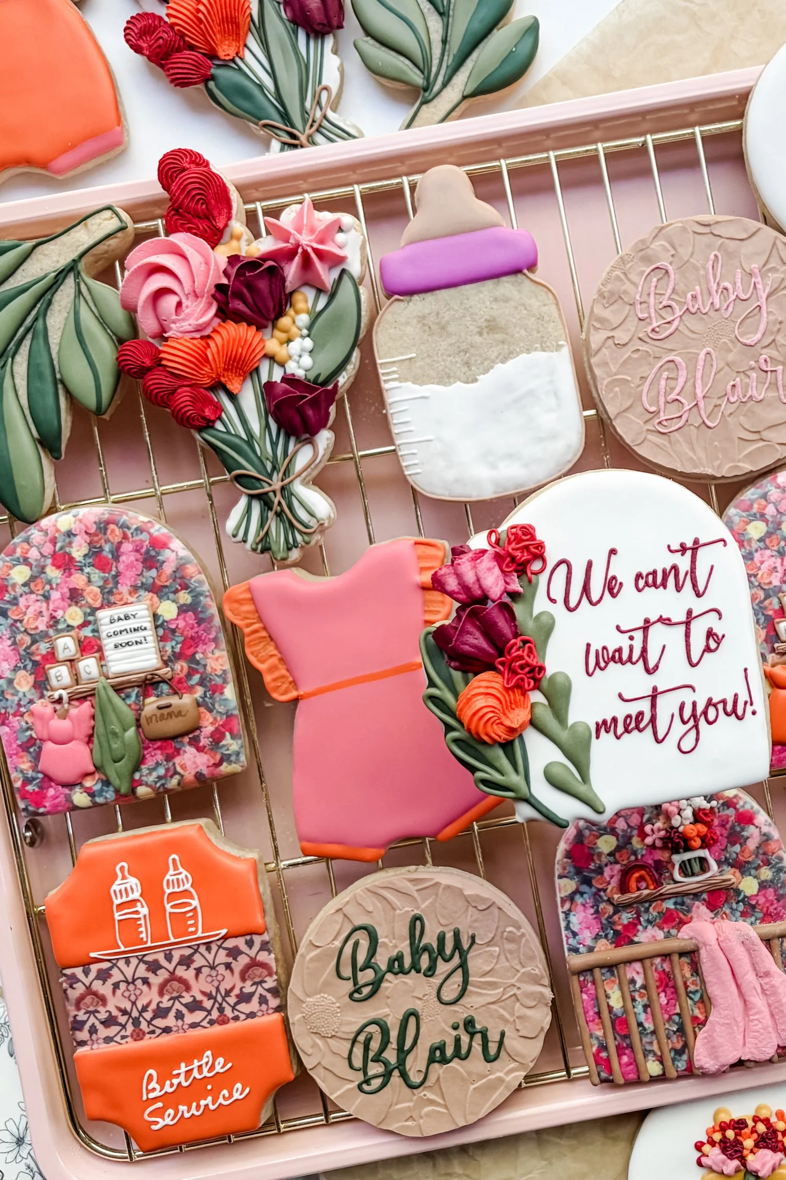 Decorative cookies shaped and decorated for a baby shower, including ones with floral designs, baby bottles, and words like 'Baby Girl' and 'Baby Blair,' displayed on a pink tray.