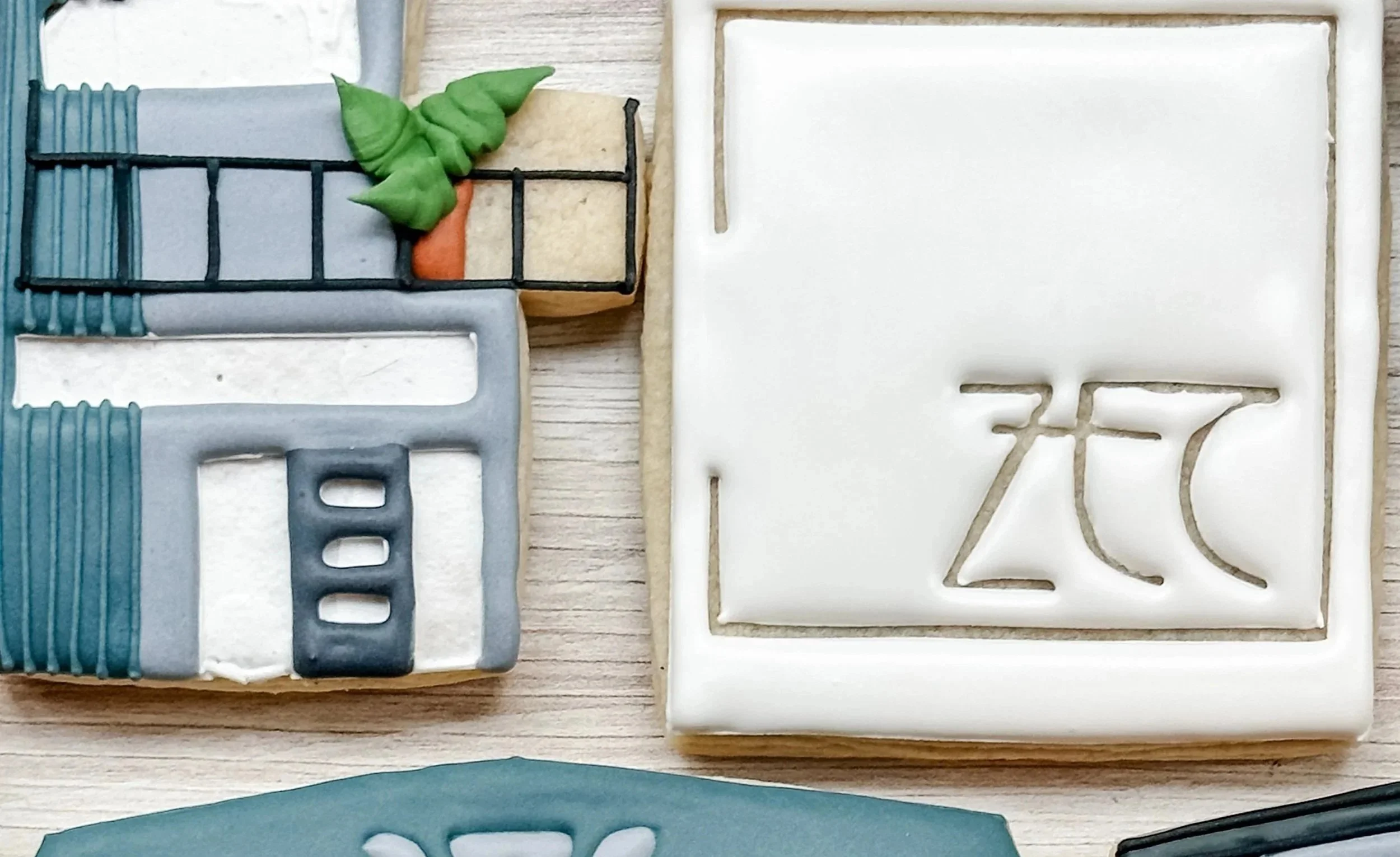 Zack Evans Construction Logo Cookies decorated with royal icing, and modern house cookies decorated with green leaves and terracotta accents, placed on a wooden surface.