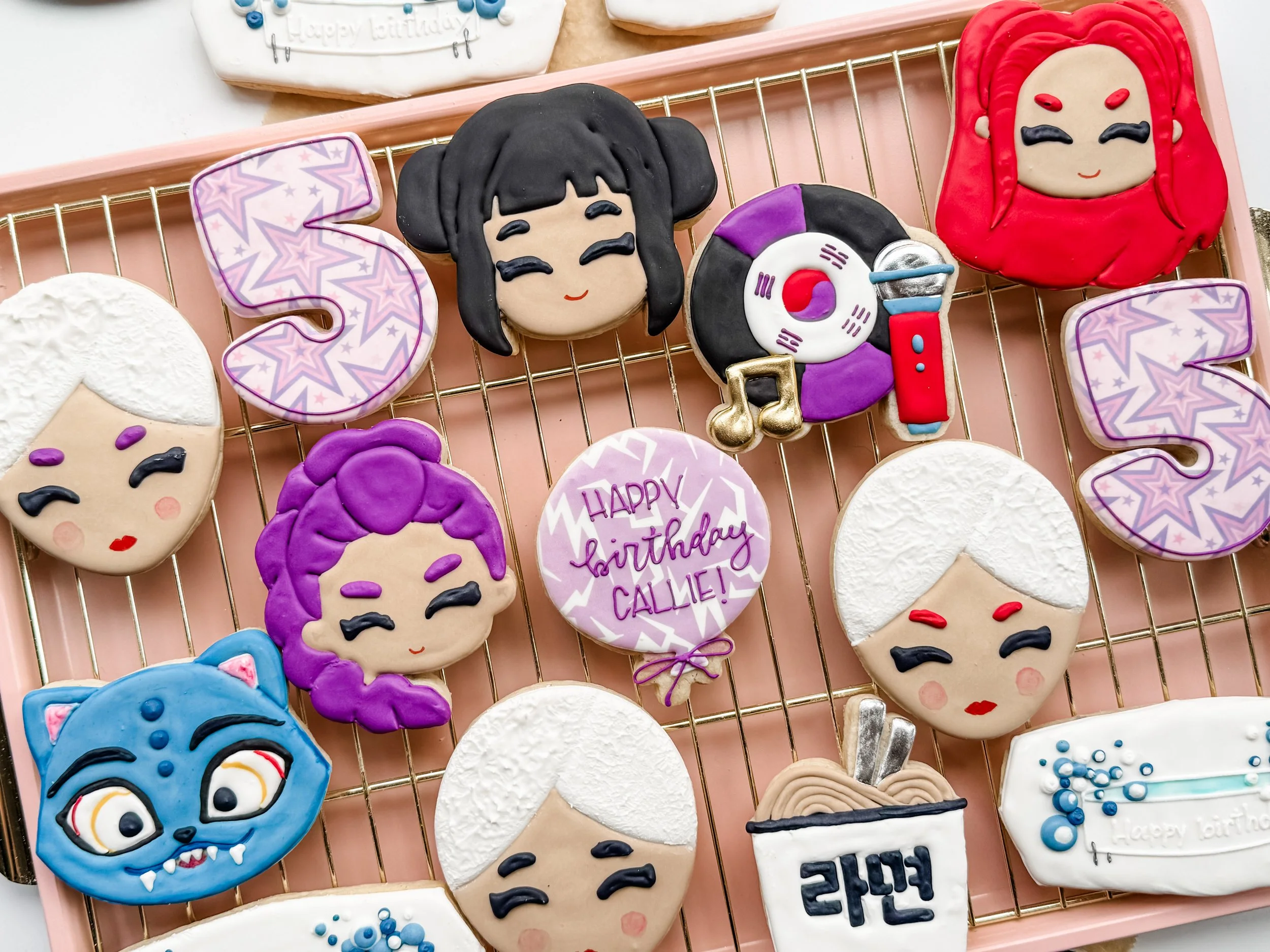 K-Pop Demon Hunters themed decorated sugar cookies with various designs, including a girl with black hair and a red hairdo, a blue cat with glasses, a vintage microphone, a purple flower, a white circle with 