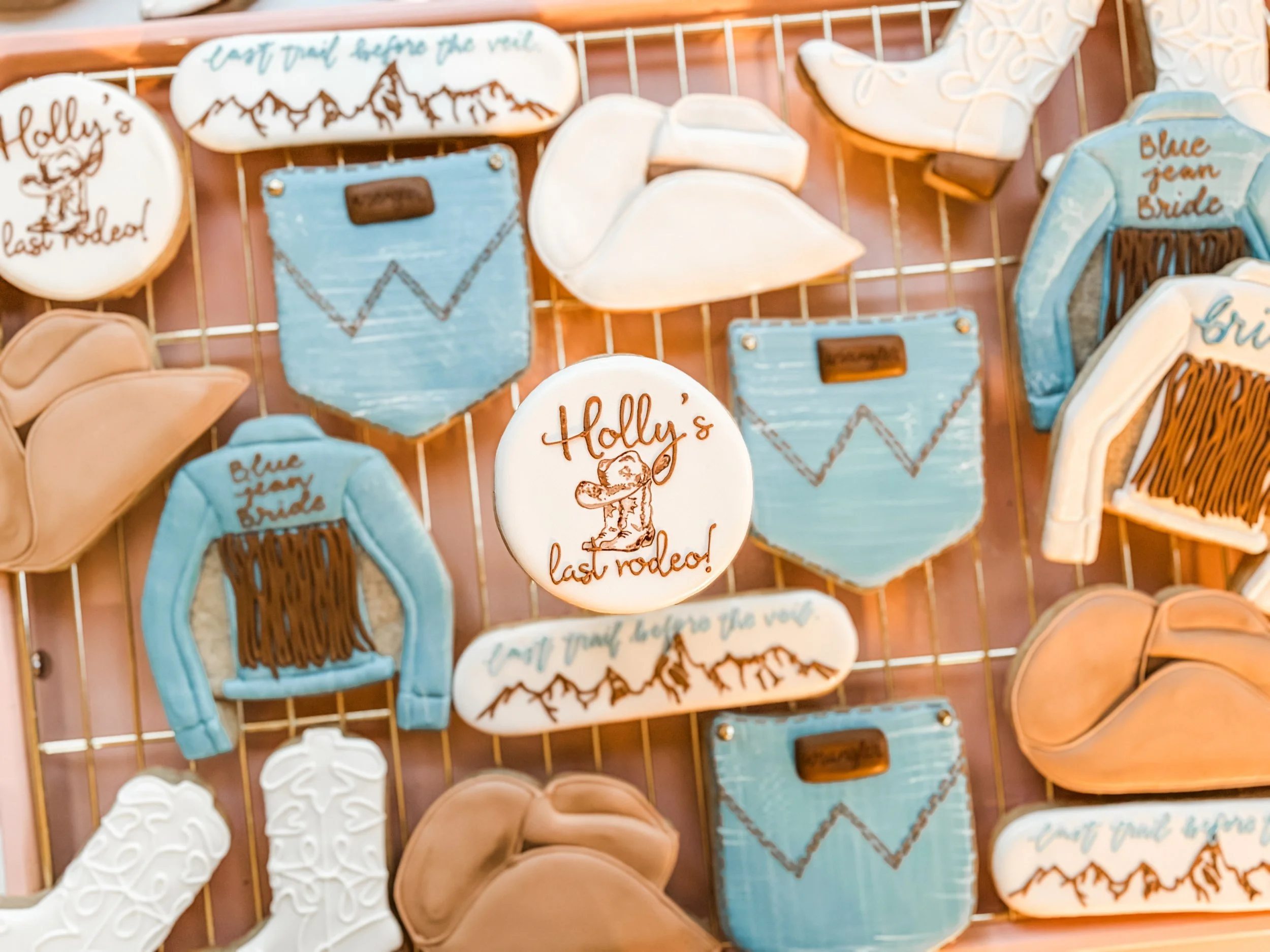 Decorative cookies themed around a mountain or travel, featuring images of mountains, a suitcase, cowgirl boots, and a jean jacket, with text references to last rodeo and a bride.