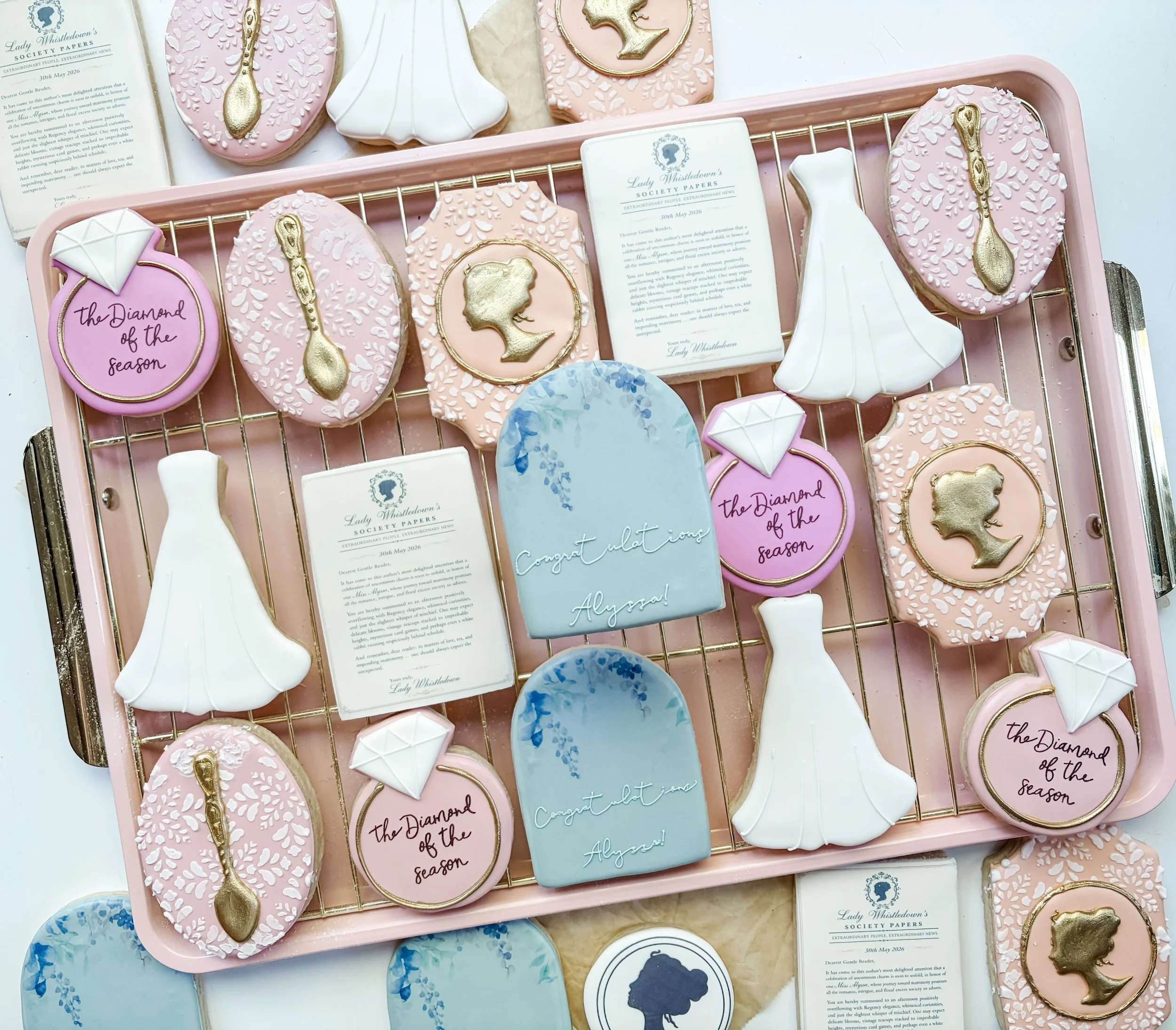 Bridgerton themed pink and white cookies decorated with gold spoons, mermaid silhouettes, and abstract designs and some cards with messages like "Congratulations Alyssa" and "the Diamond of the season" in a pink box.