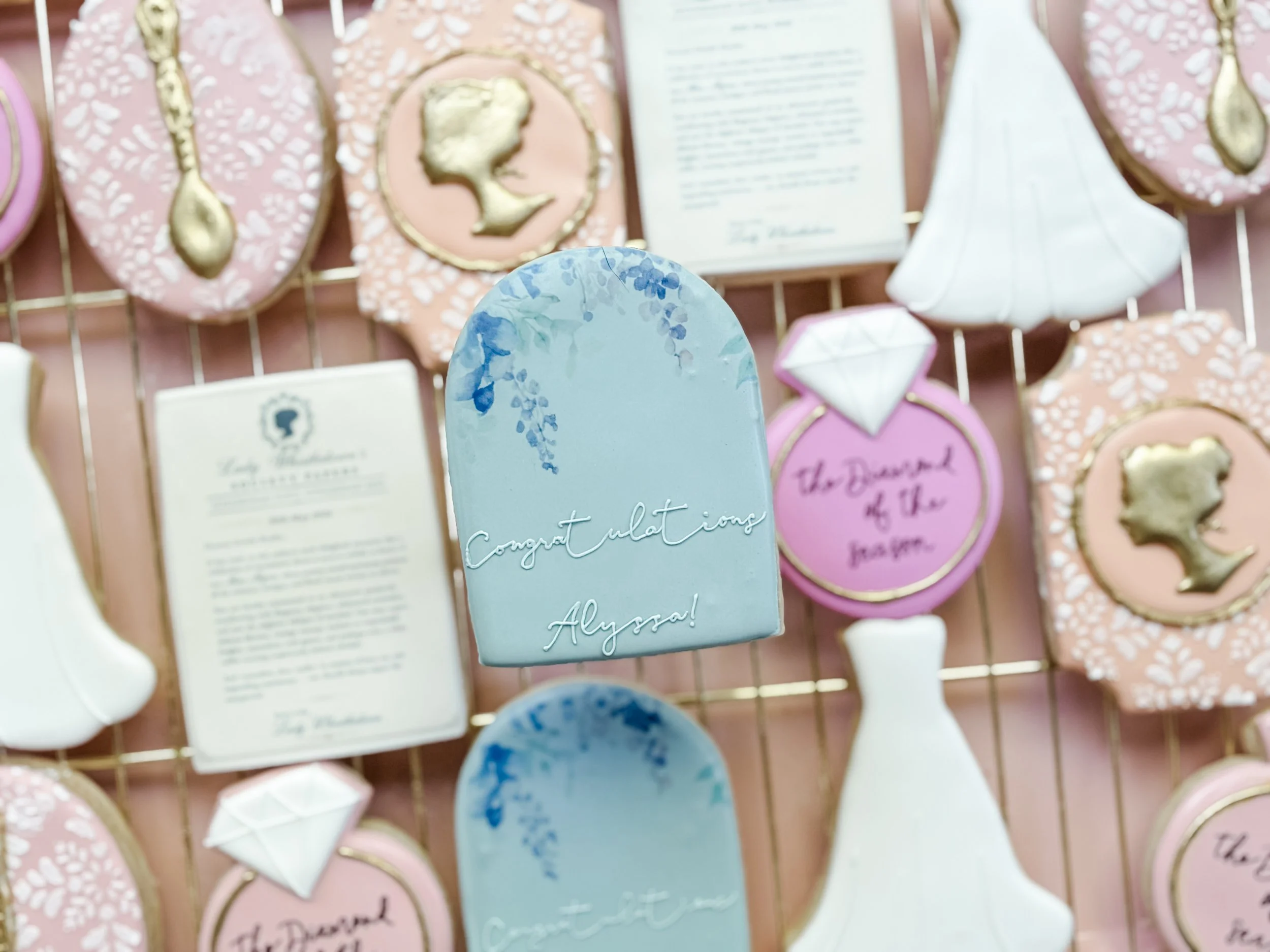 Display of various decorated cookies with pastel colors, floral patterns, and gold accents, along with handwritten notes and a framed profile silhouette in the background.