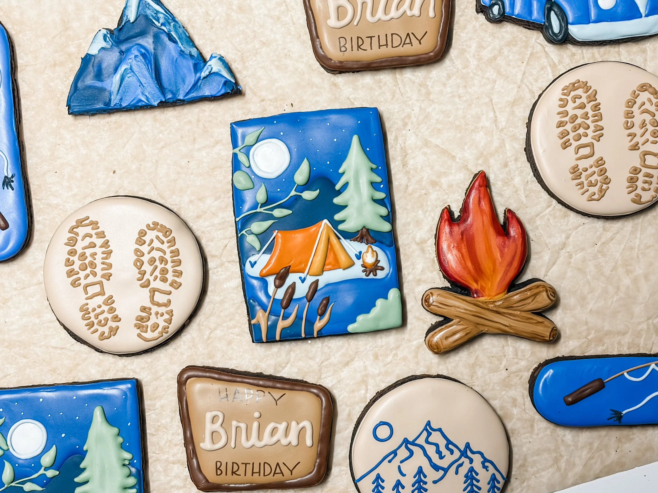 Decorated cookies with outdoors and birthday themes, including trees, a tent, campfire, mountains, and words such as 'Happy Birthday' and 'Dream'.
