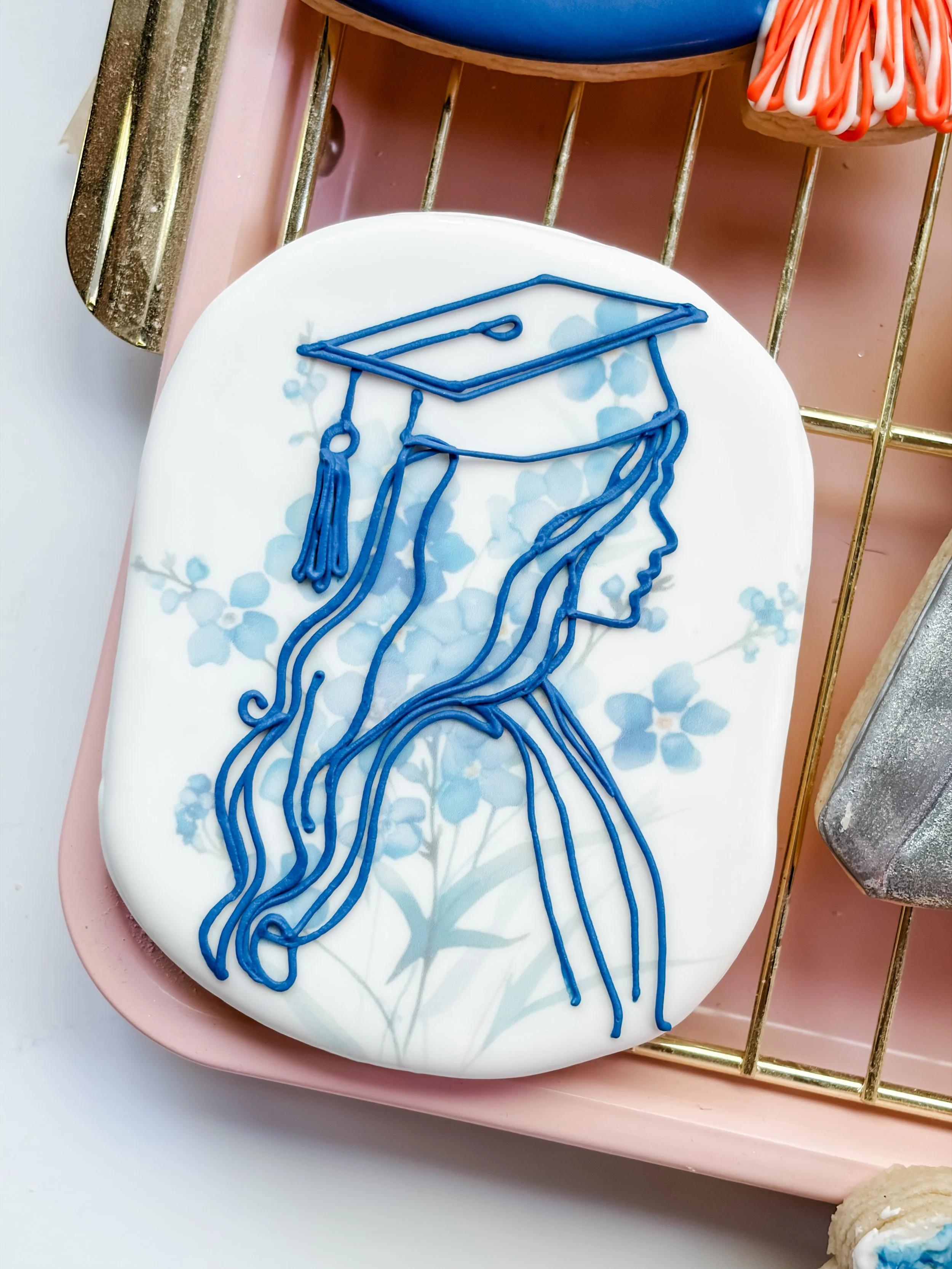 A white plate with a blue outline of a woman wearing a graduation cap drawn with icing. The plate is on a pink tray with a gold wire rack underneath it, holding decorated cookies.