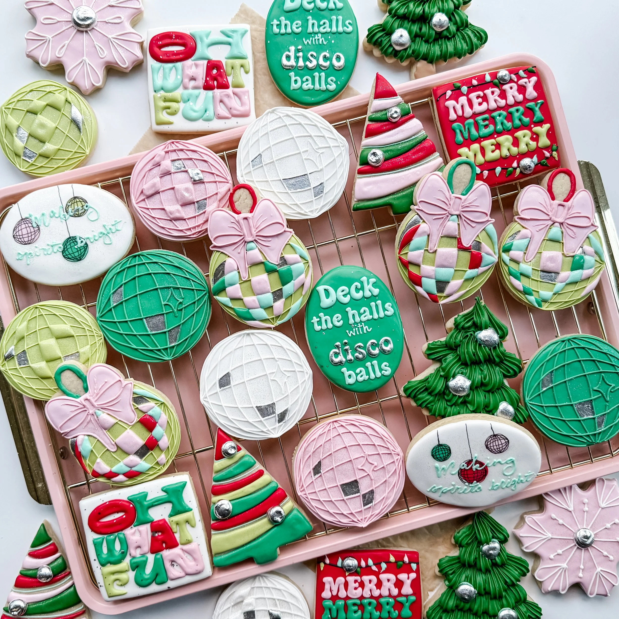 Assorted decorated Christmas cookies on a pink tray, including a Christmas tree, ornaments, bows, a Santa hat, and holiday messages like "deck the halls with disco balls" and "merry merry" in bright colors and festive designs.