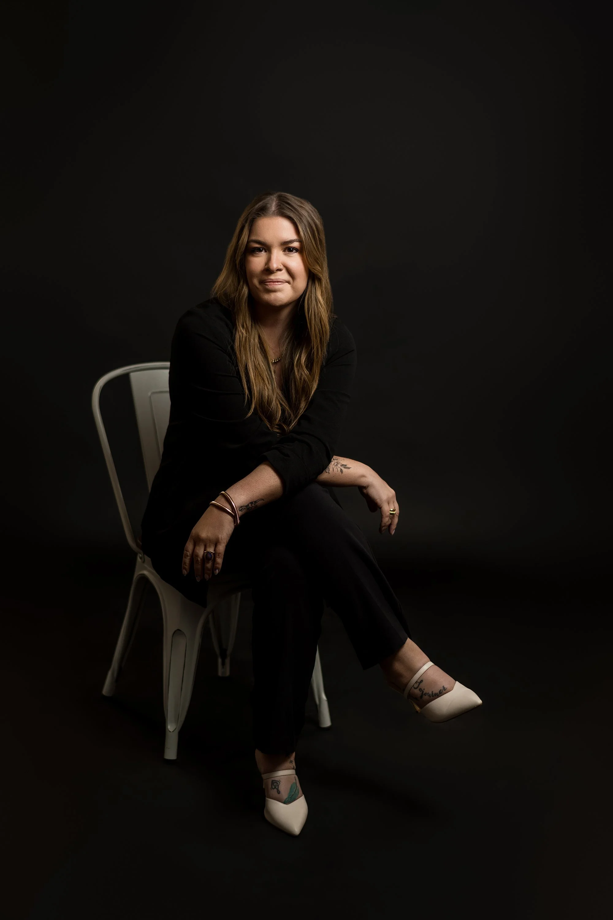 Portrait of a woman with long brown hair sitting on a white chair against a black background, wearing a black outfit. This is imagery for her SEO Optimized Squarespace Website Build.