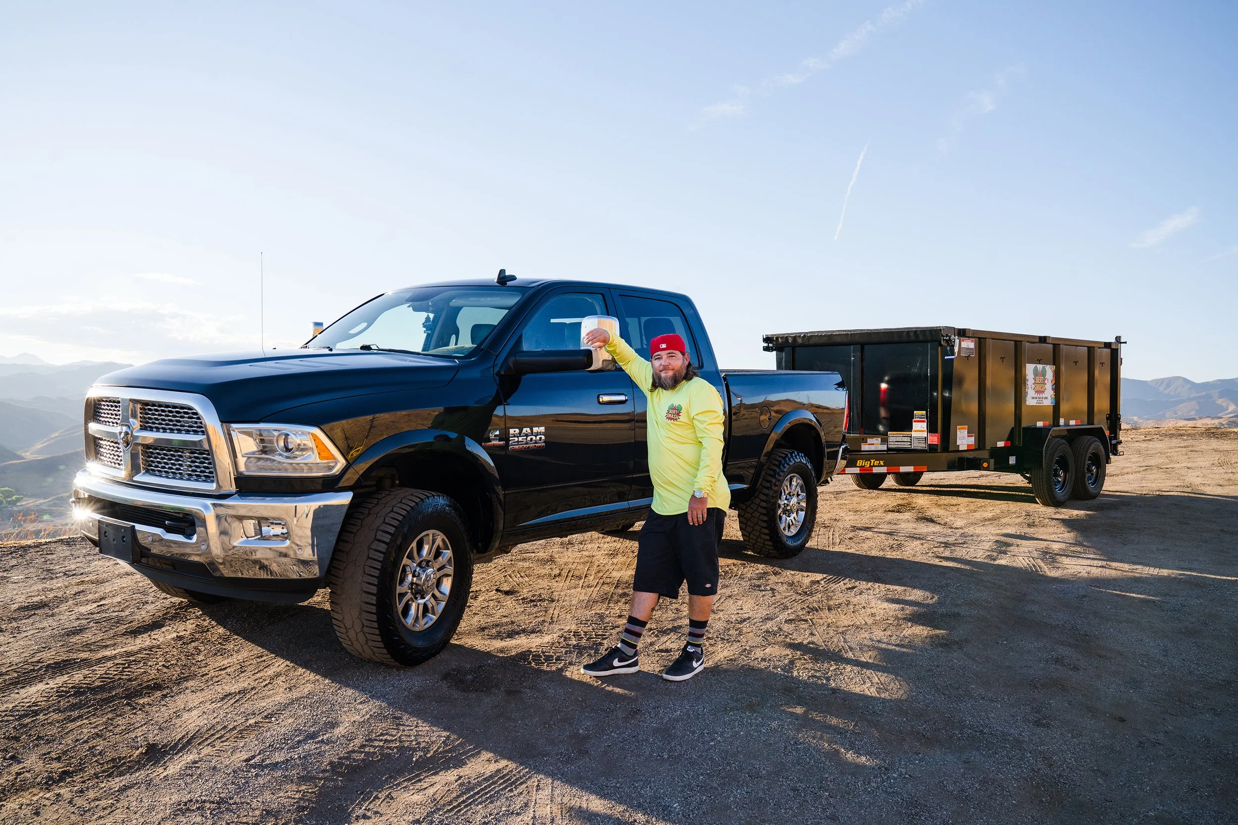 A man with a beard, wearing a red cap, yellow shirt, and black shorts, is standing next to a black RAM 2500 pickup truck. He is using a Small Business Systems Consultant to startup his small business.