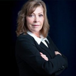 Headshot of Meribeth, a client of The Simple Mouse, smiling softly in a black blazer and white collared shirt with her arms crossed against a dark background.