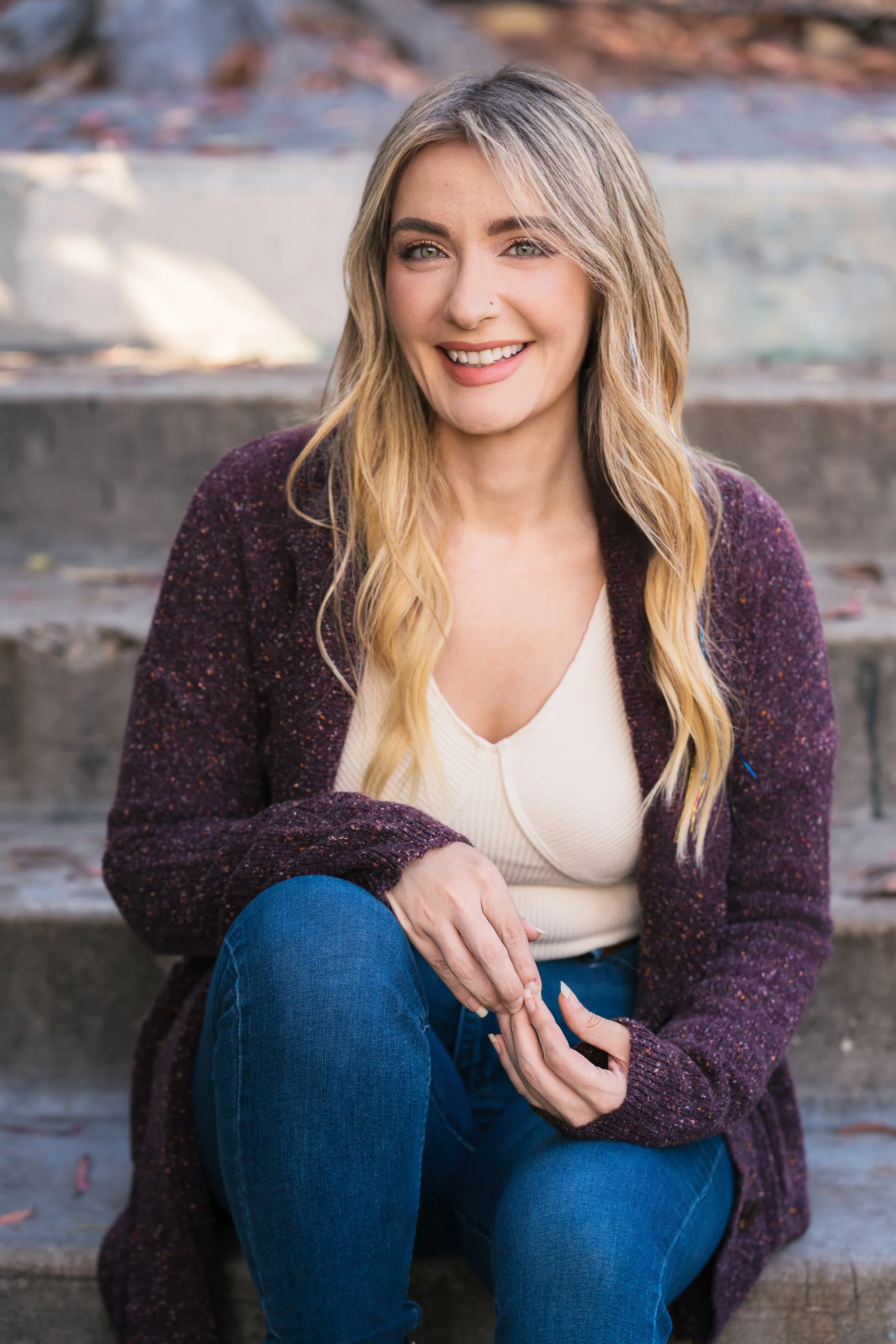 A woman with long blonde hair sitting on outdoor steps, smiling and looking at the camera. She is wearing a cream top, a purple cardigan, and blue jeans for an example of professional small business launch services.