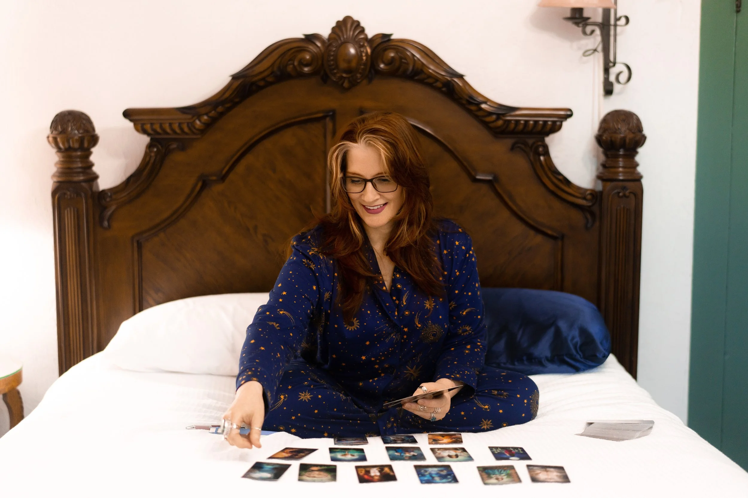 A woman with red hair, glasses, and a blue star-patterned pajama set sits on a bed, smiling, as she sorts through tarot cards laid out in front of her, prepping for her small business launch services with The Simple Mouse.