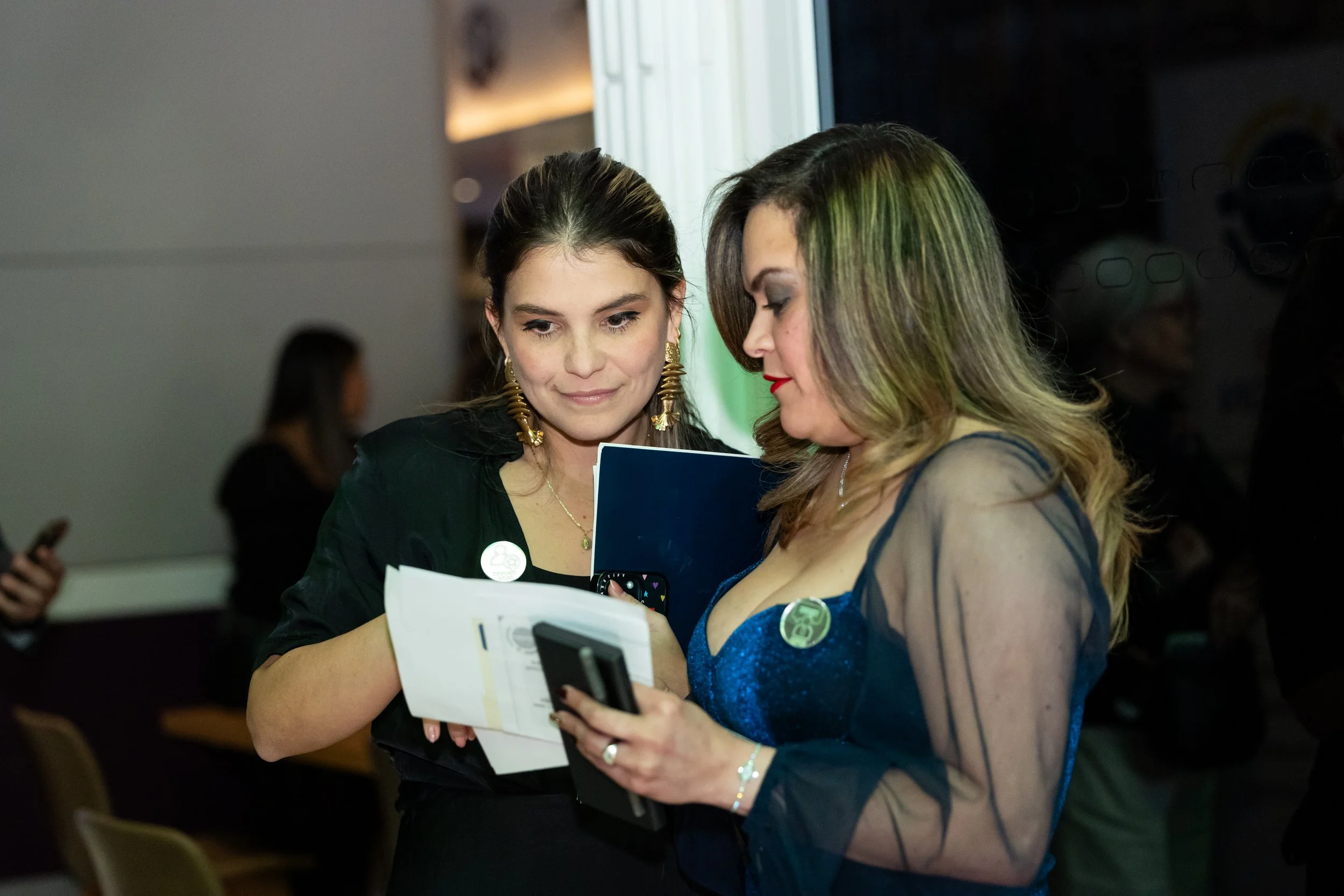 Two women are looking at a smartphone and papers together in an indoor setting, possibly at an event or conference.