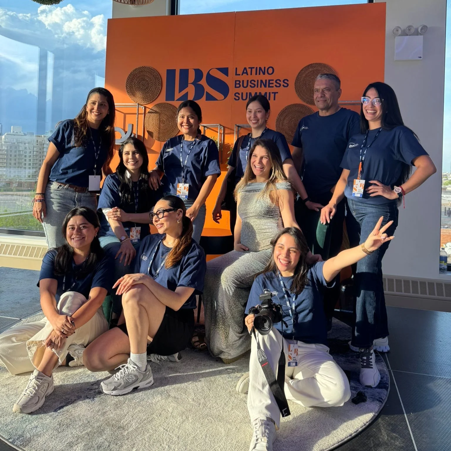 Group of professionals attending the Latino Business Summit posing for a photo on a stage with a large orange backdrop that reads LBS Latino Business Summit, with cityscape visible through the window.