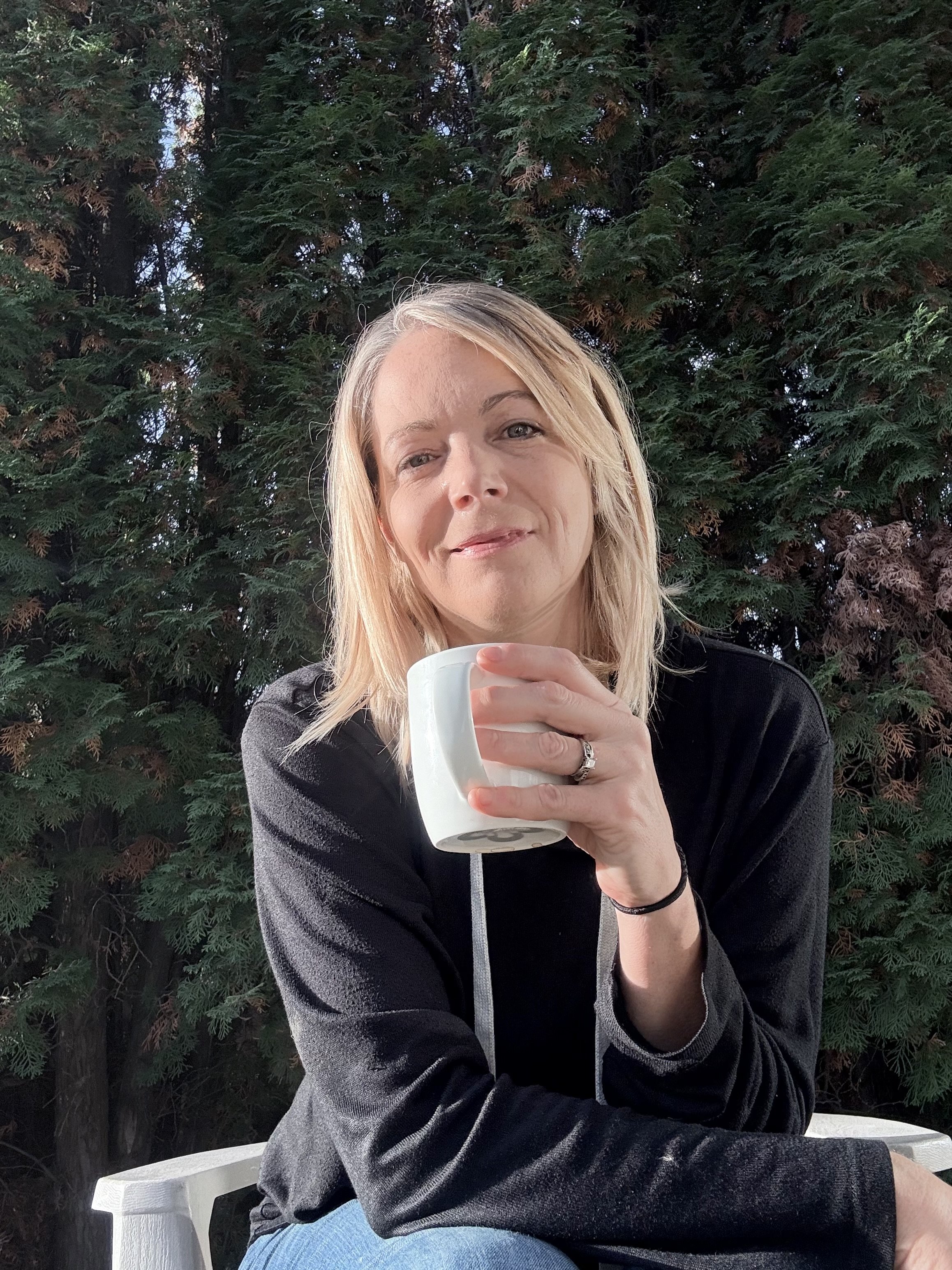 A woman with blonde hair sitting outdoors, holding a white mug, smiling slightly, with a large green tree in the background.