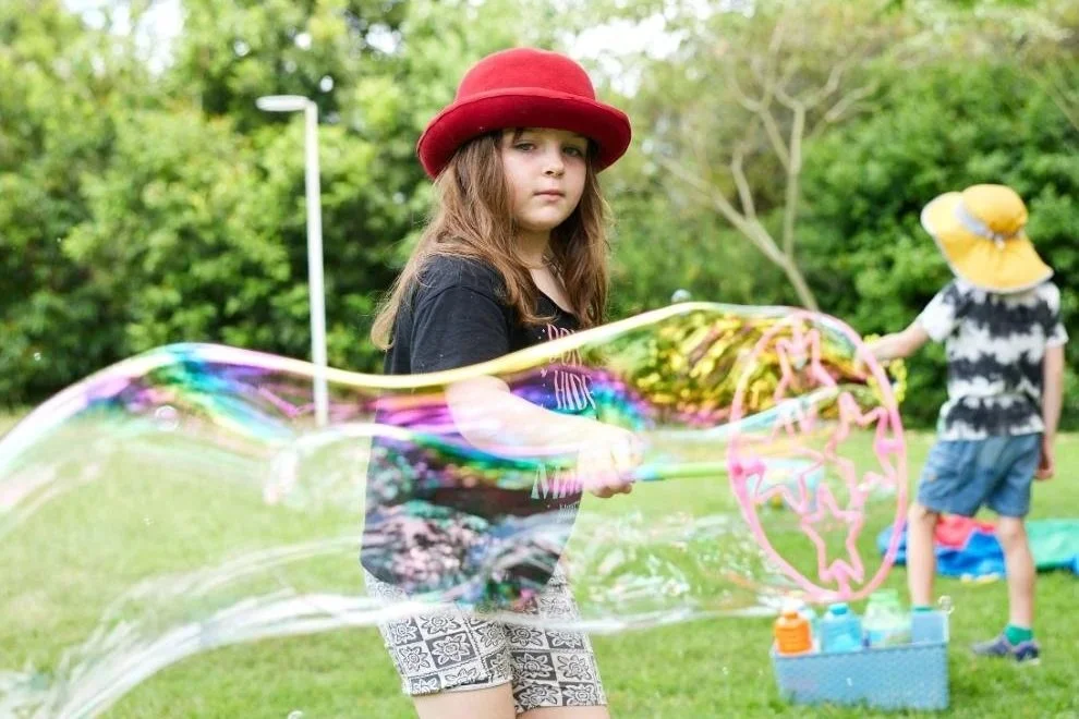 A young girl with a red hat making a large soap bubble outdoors, with other kids in the background, surrounded by green trees and grass.