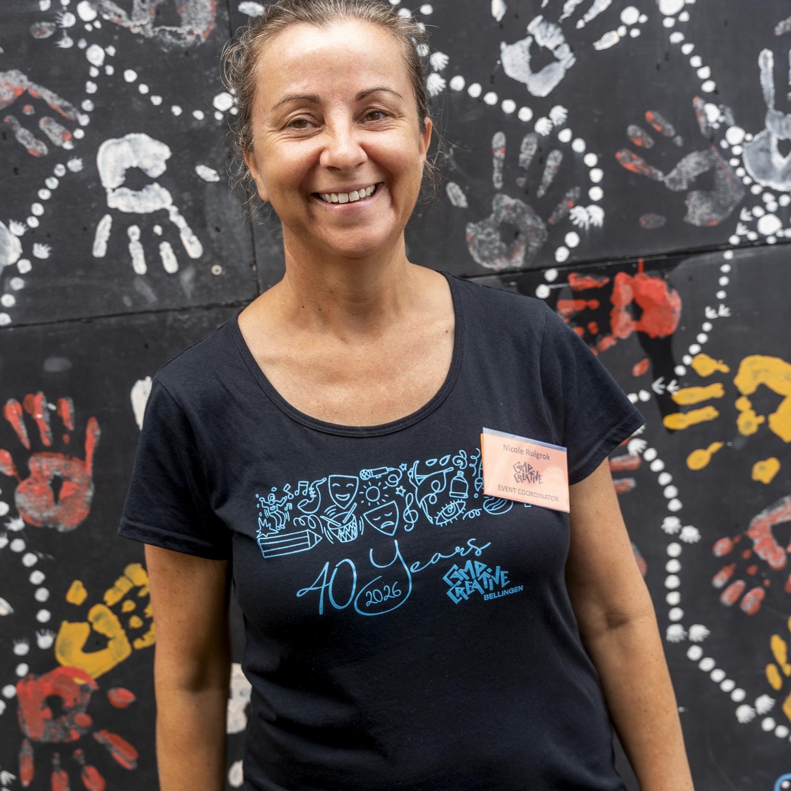 Smiling woman wearing a black T-shirt with a design celebrating 40 years of GMA Creative in 2026, standing in front of a black background with colorful handprints and doodles.