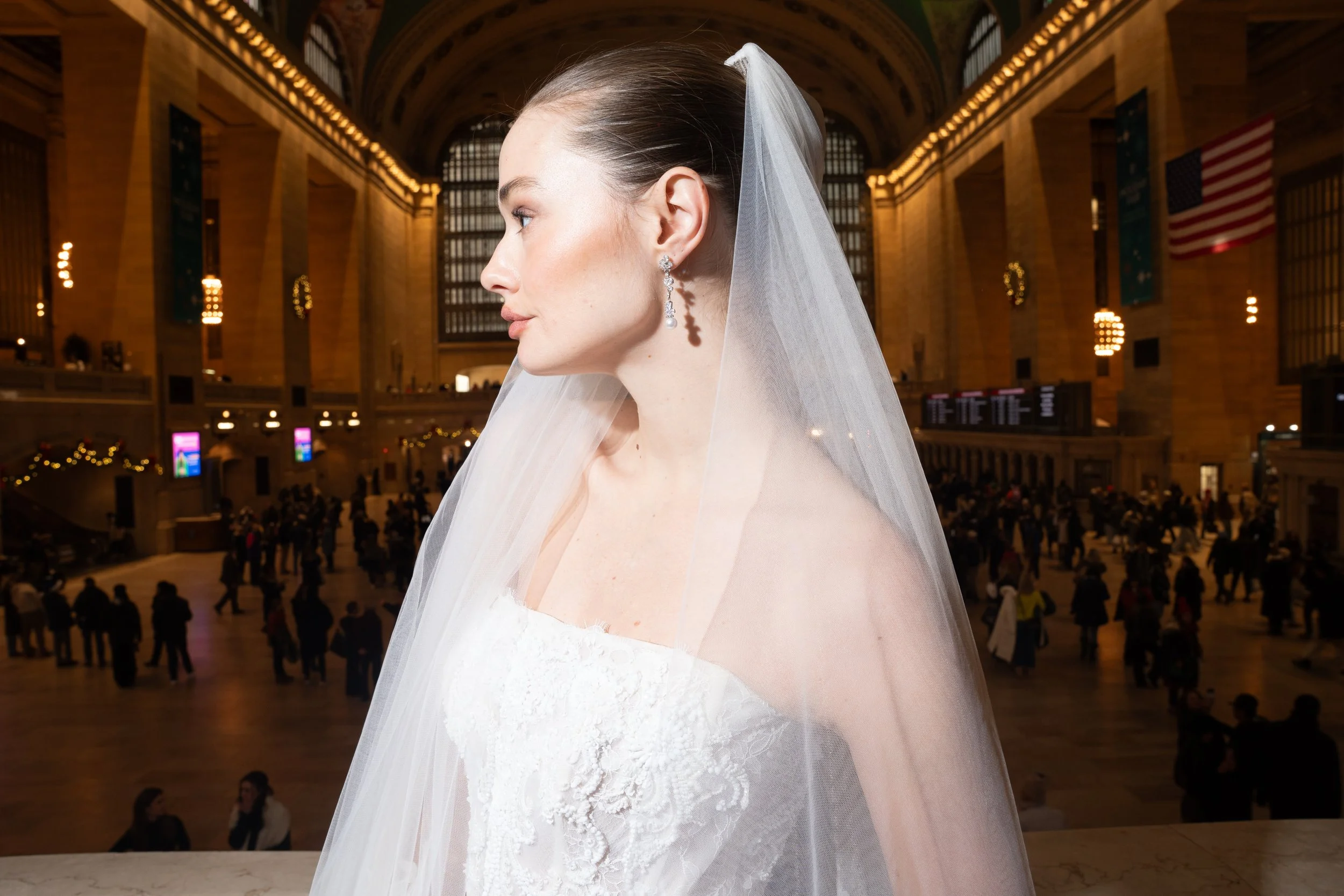 A bride in a white wedding dress and veil inside a train station with large windows, flags, and people in the background.