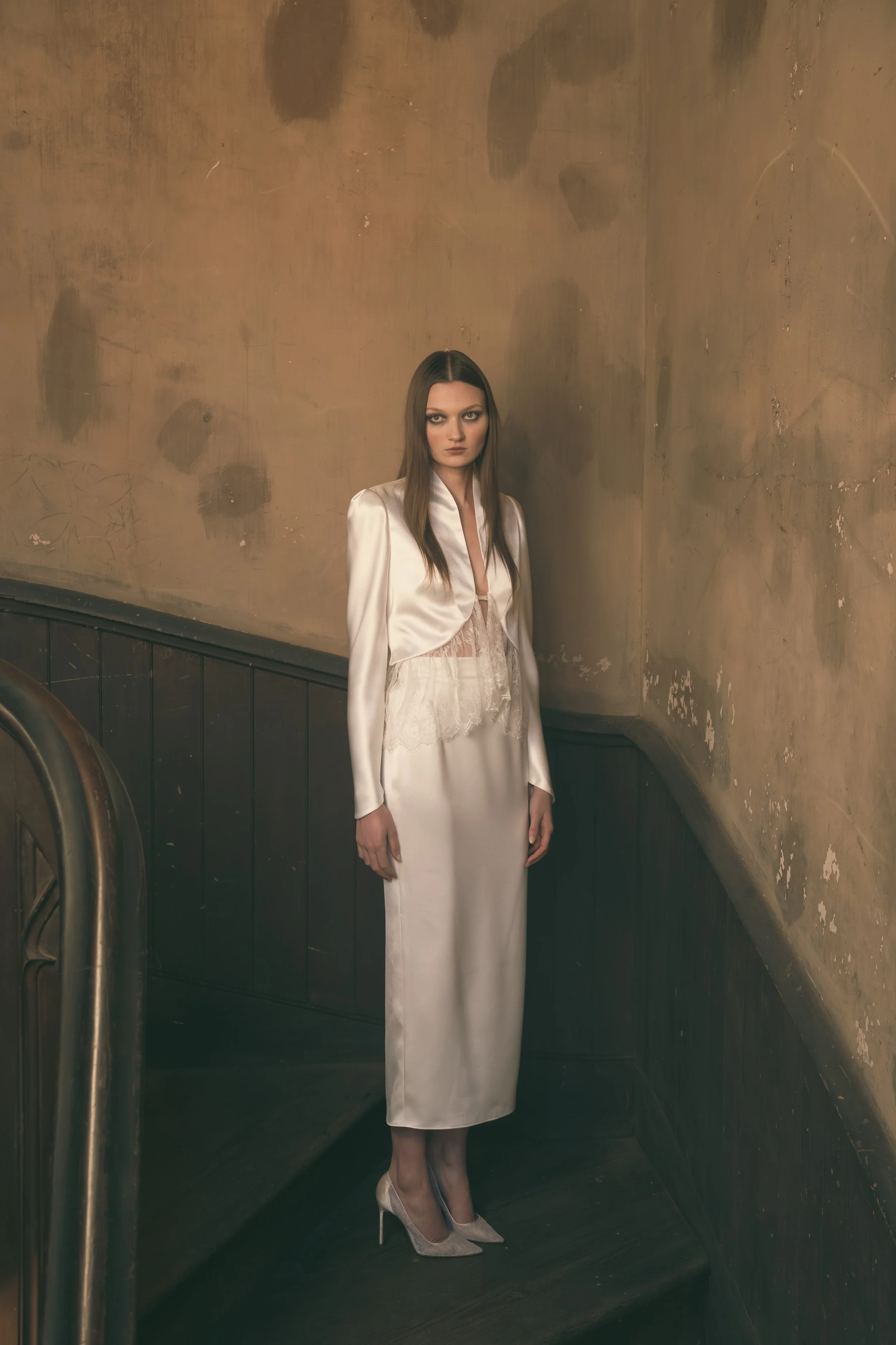A young woman with long brown hair wearing a silky white jacket, lace top, satin skirt, and high heels standing on a staircase in a dimly lit, slightly worn space with textured beige walls.