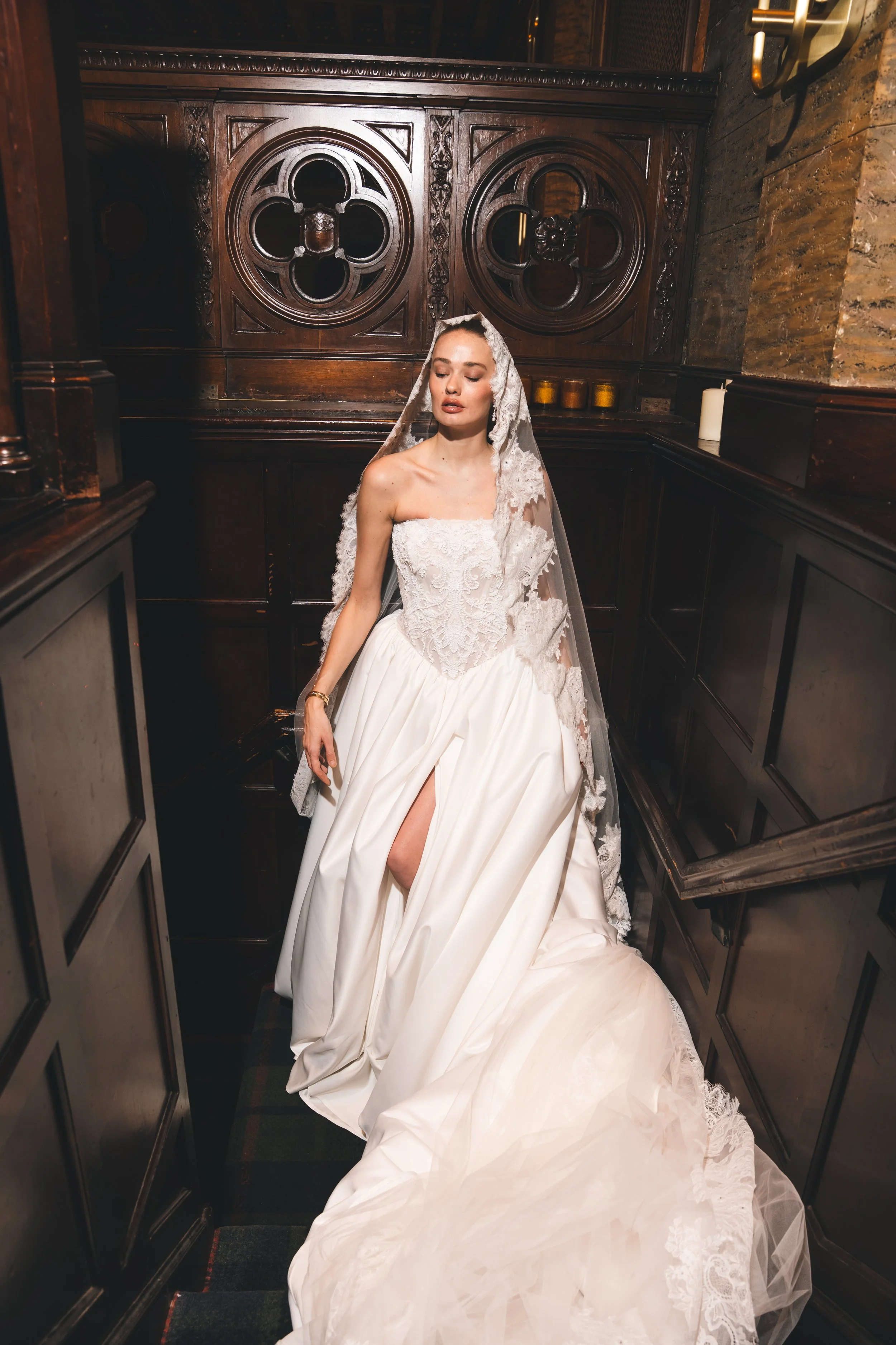 A bride in a white wedding dress and lace veil stands on a staircase in a wooden-paneled, dimly lit room with dark wood accents and a stone wall.