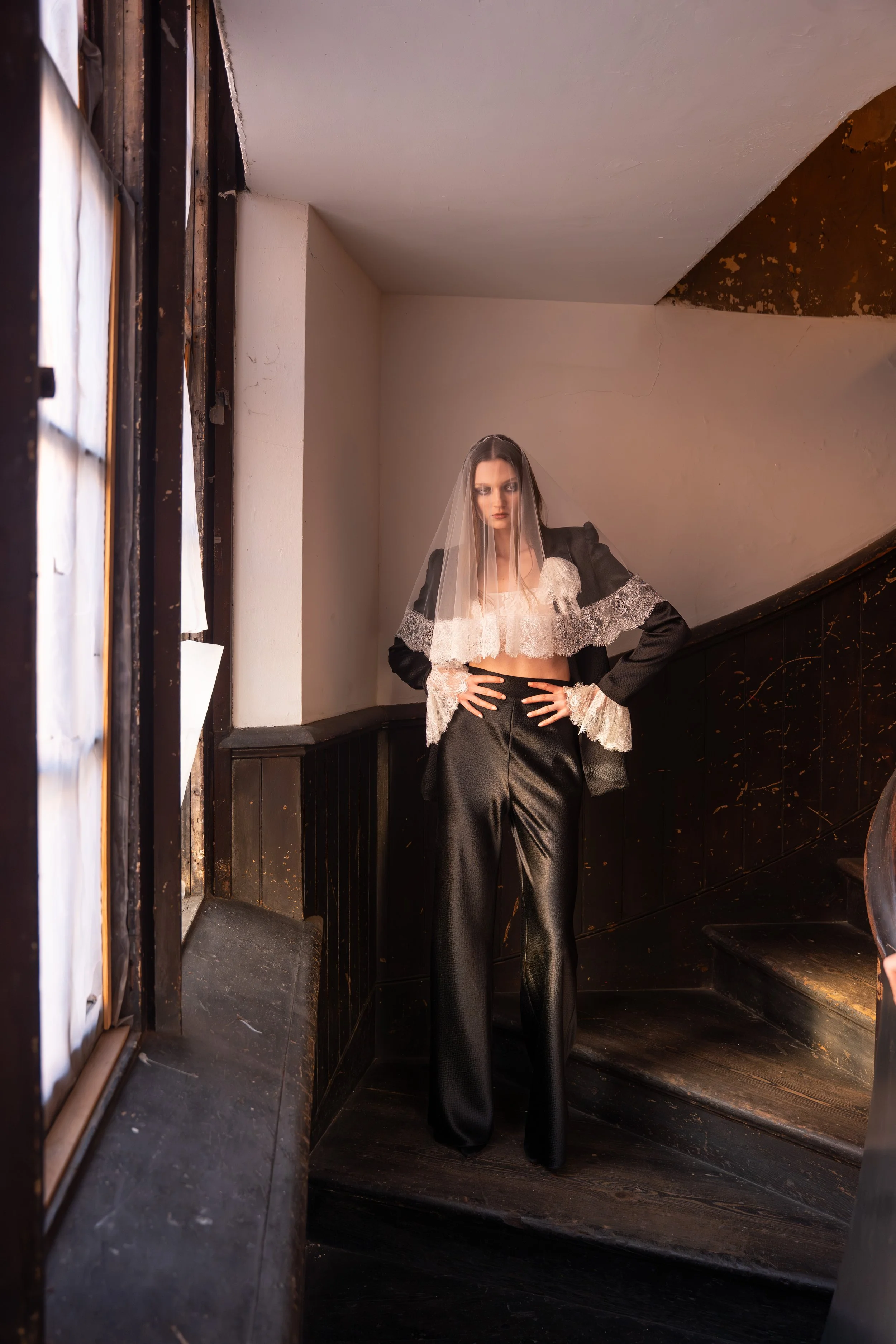 Woman standing on a staircase in a black satin pantsuit with lace accents, sheer veil, and lace cropped top, posing indoors with one hand on her hip.