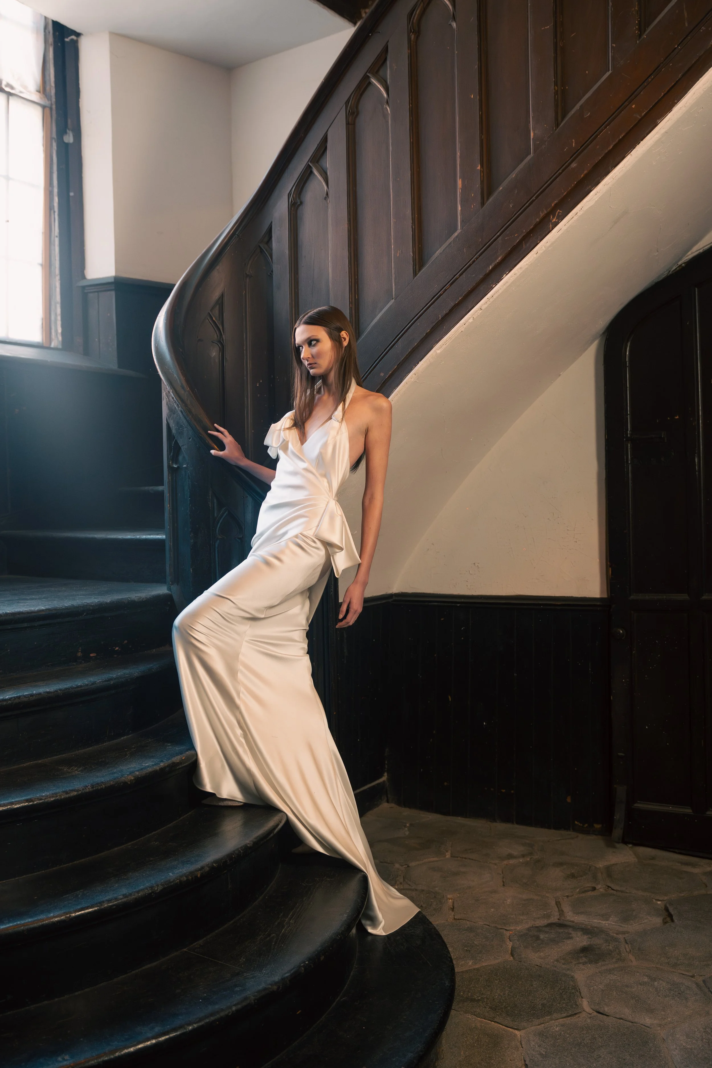 A woman in a long, silky white dress standing on a curved, black wooden staircase in a historic building with large windows.
