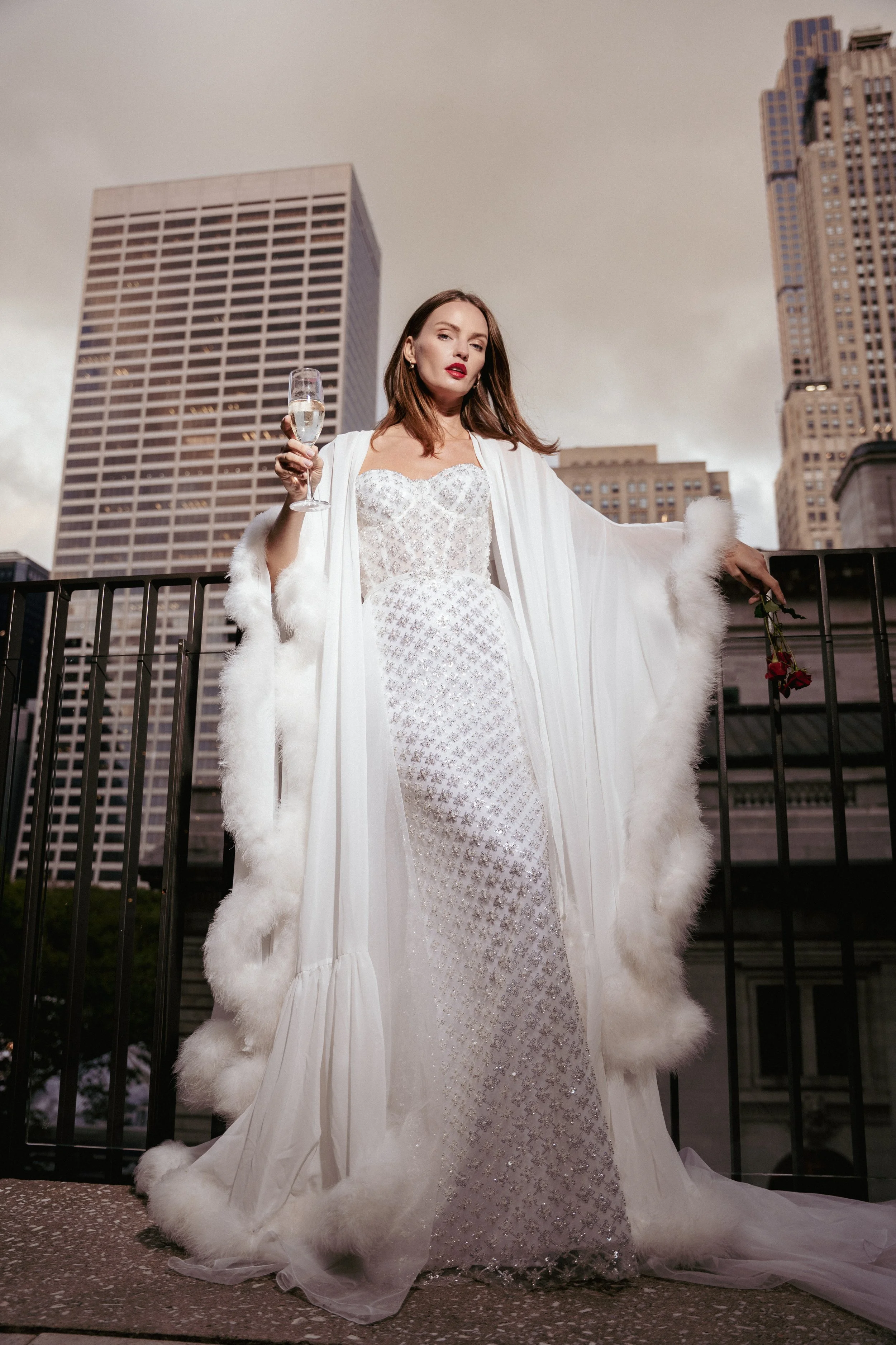 A woman in a white, elegant dress and long flowing robe with fur trim standing on a balcony in an urban setting, holding a glass of champagne and a single red rose.
