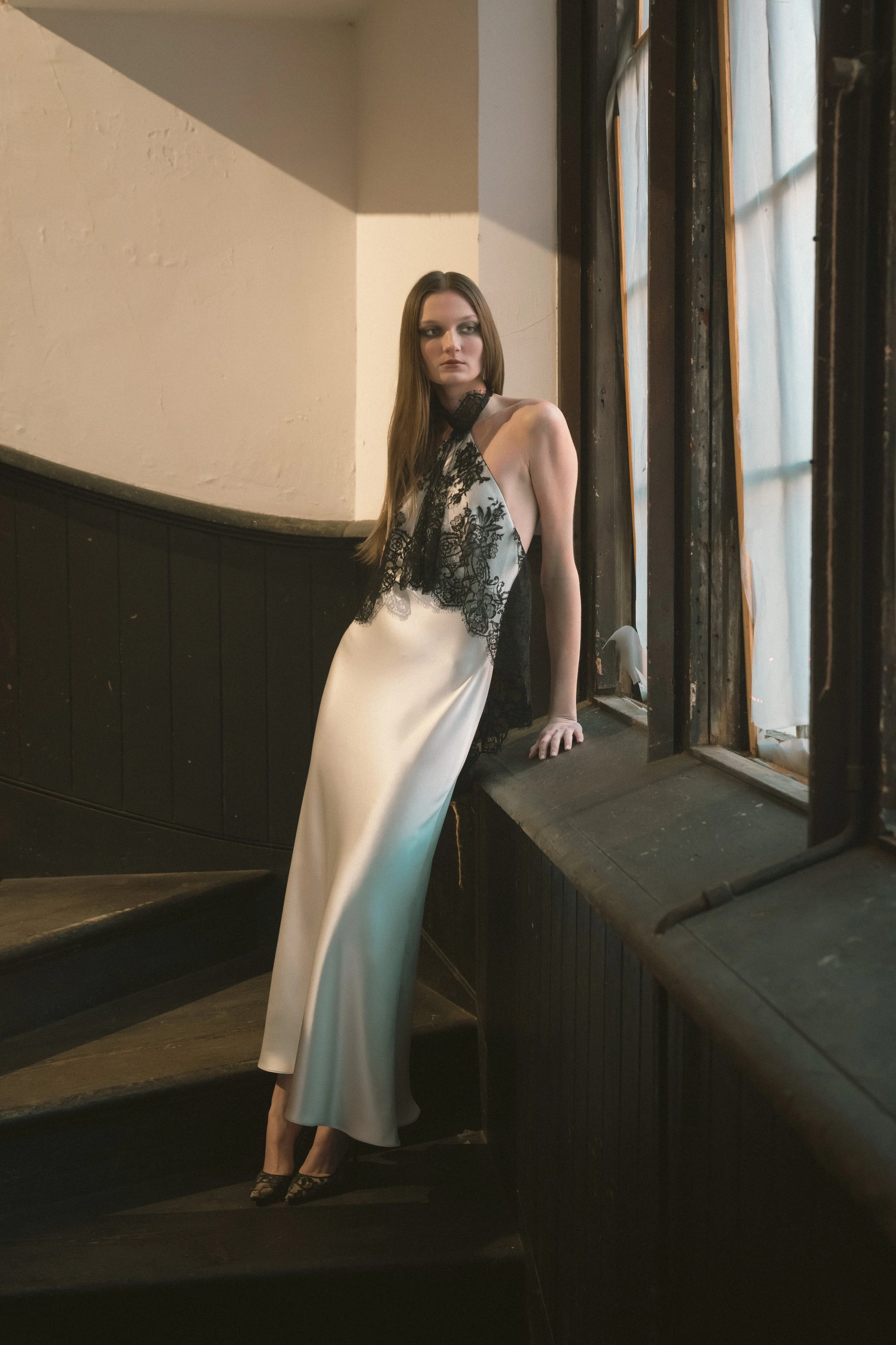A woman in a satin dress with black lace details leaning against a windowsill in a dimly lit interior.