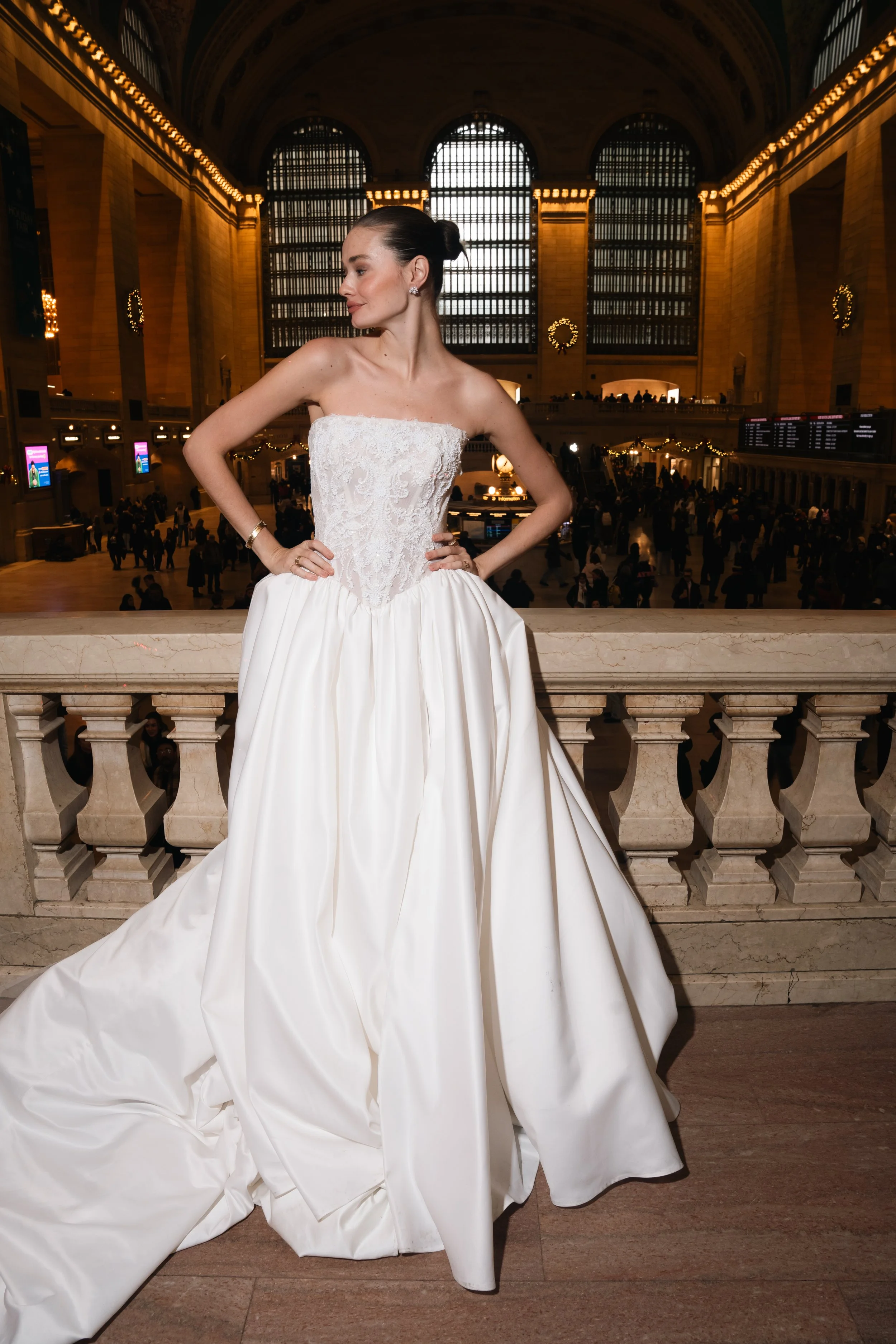 A woman in a white strapless wedding gown with a lace bodice and voluminous satin skirt standing inside a grand train station with high arched windows and a marble railing.