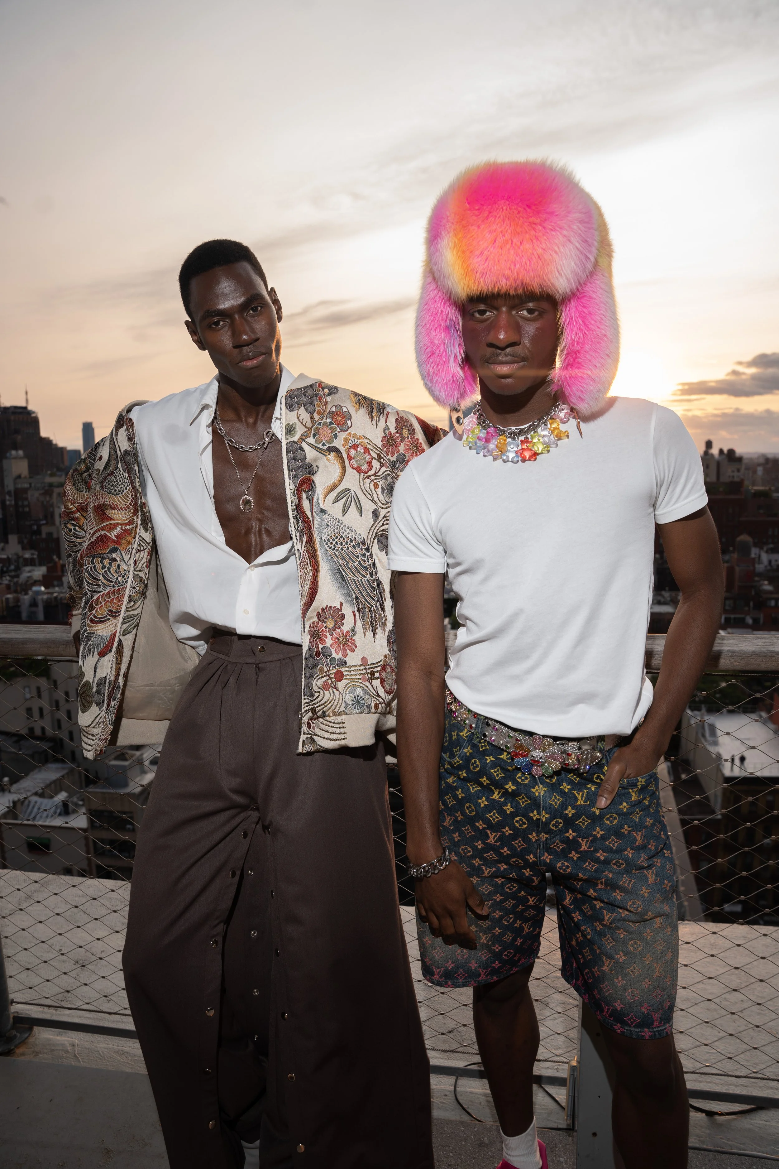Two young men standing on a rooftop during sunset, dressed in fashionable clothing with one wearing a colorful furry hat and jewelry, and the other wearing a patterned jacket and high-waisted pants.