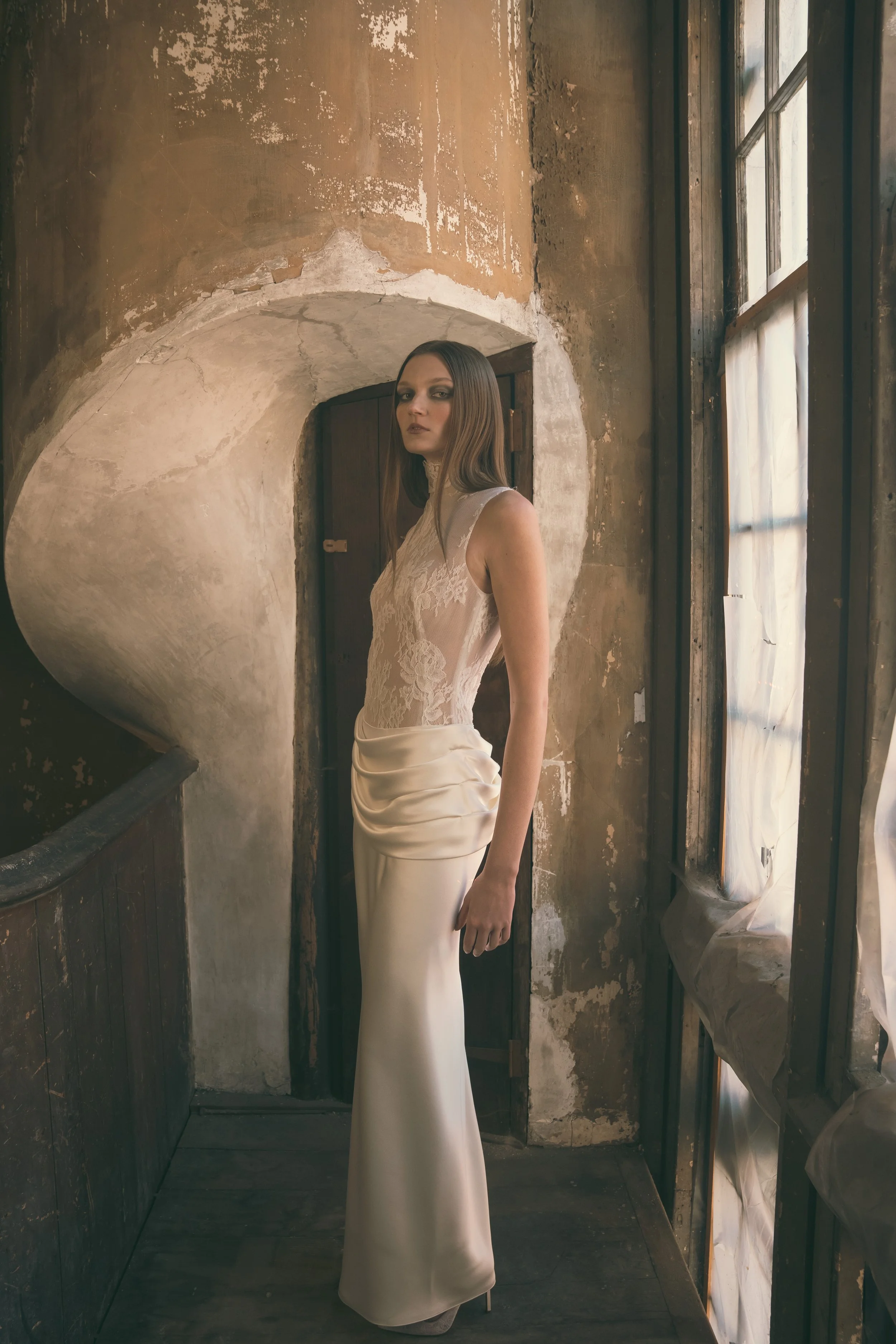 A woman in an elegant lace and satin gown stands in a rustic, weathered interior next to a window with sheer curtains.