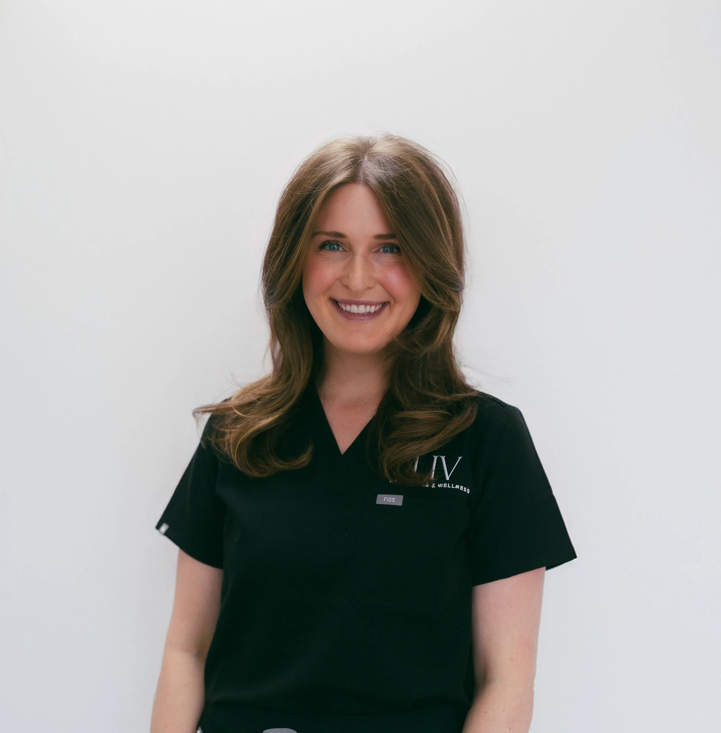A smiling woman with shoulder-length brown hair wearing a black medical uniform with a logo on the left. She stands against a plain, light-colored background.