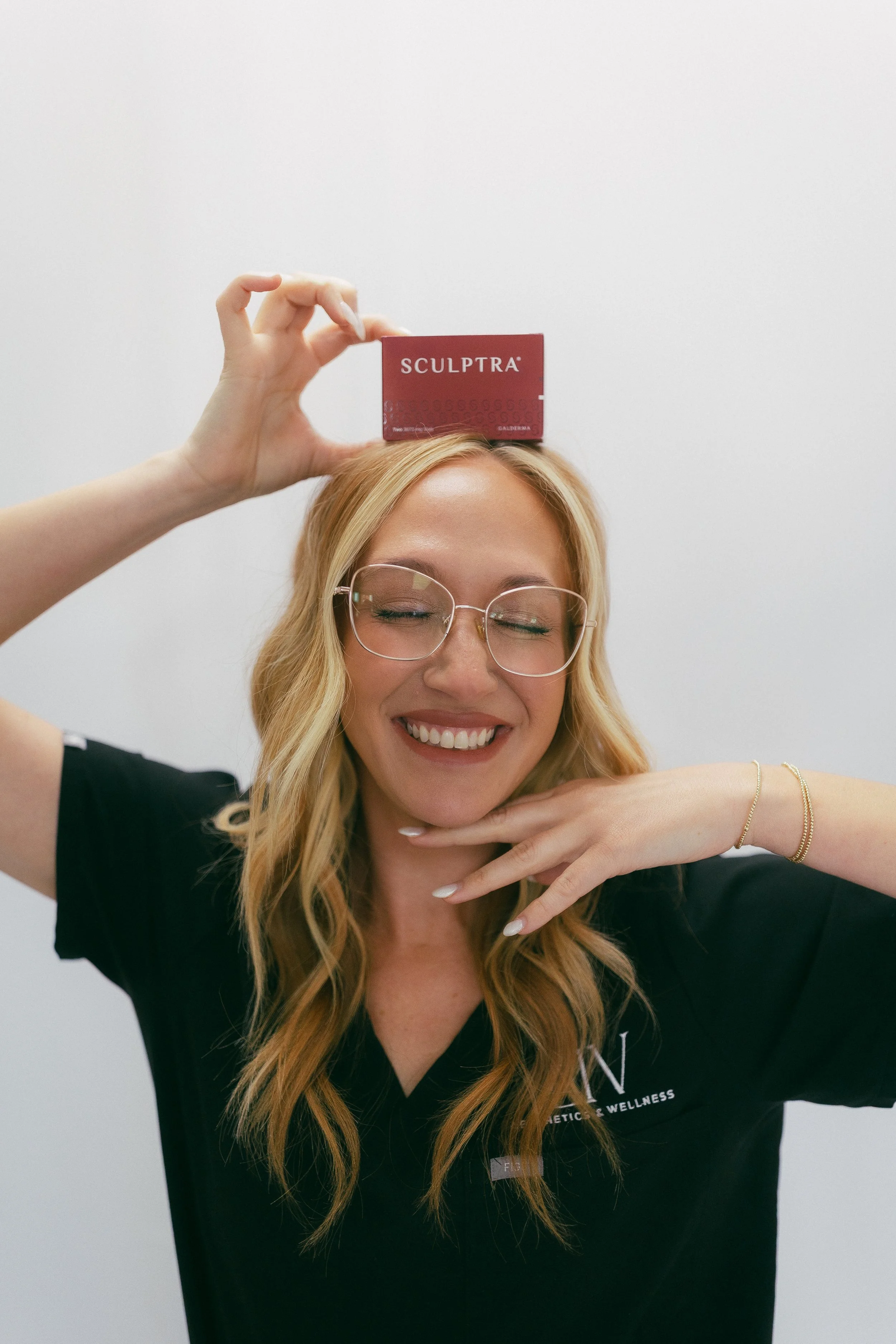 A woman with blonde hair and glasses smiling with her eyes closed, holding a red box labeled 'SCULPTRA' above her head.