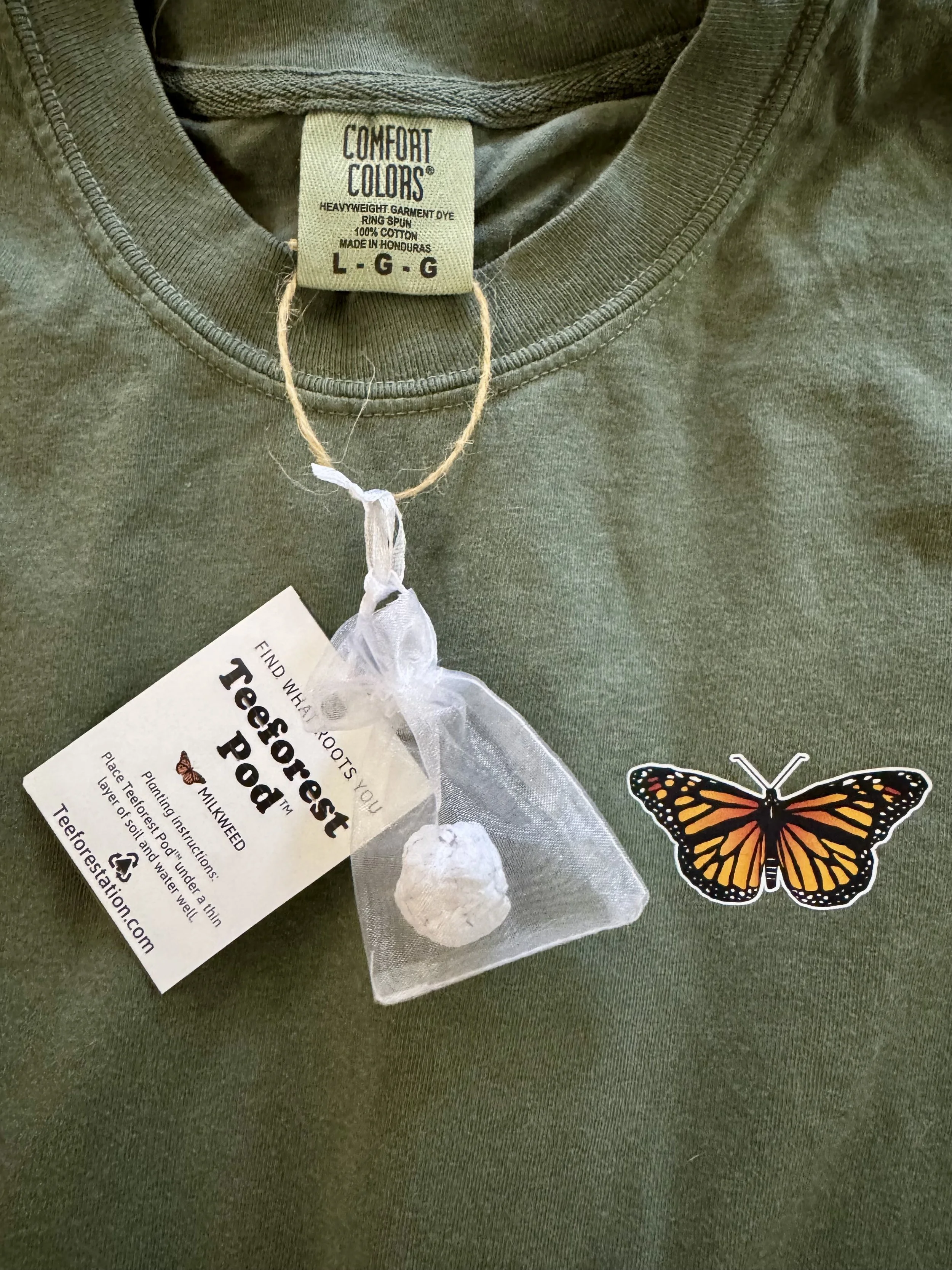 Folded Monarch Butterfly t-shirt by Evergreen Drive with a Teeforest Pod seed paper tag, garment label, and monarch butterfly logo.