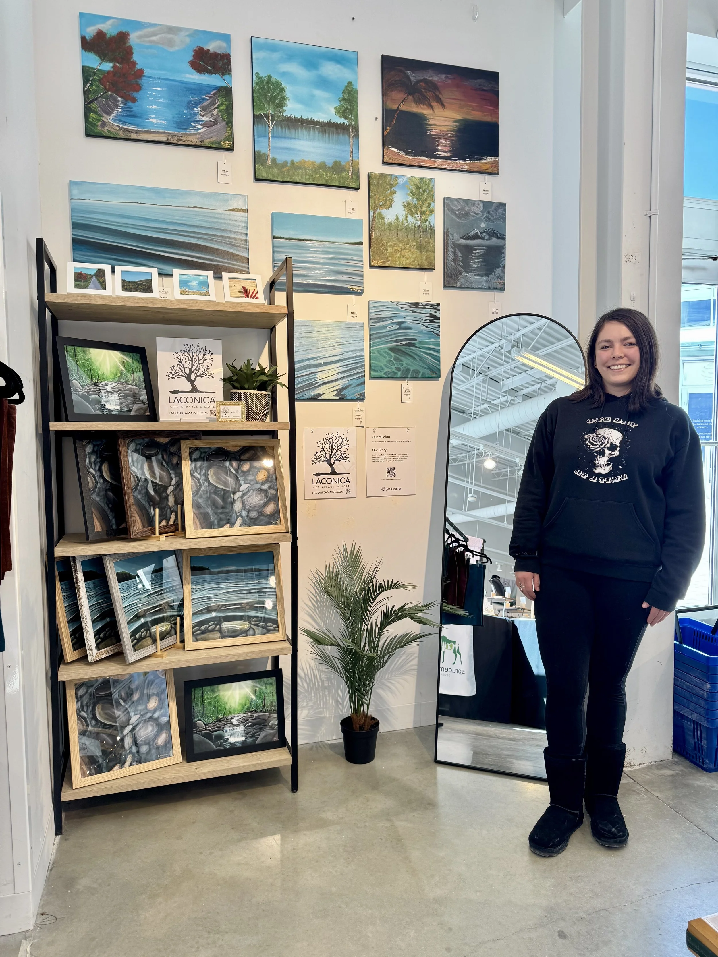 An art gallery with landscape paintings and a woman standing next to a mirror. paintings depict lakes, trees, and sunsets. The woman has shoulder-length brown hair, is smiling, and wears a black hoodie with a skull design, black pants, and boots.