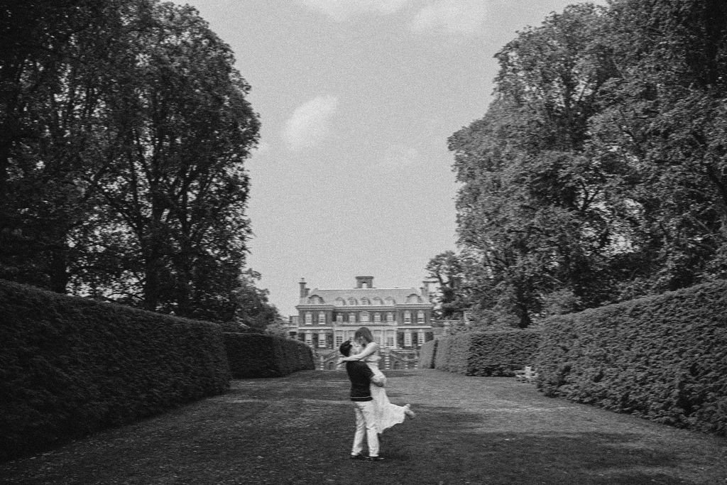 A couple dancing on a lawn in front of a large, historic mansion, with tall trees and bushes lining the sides.