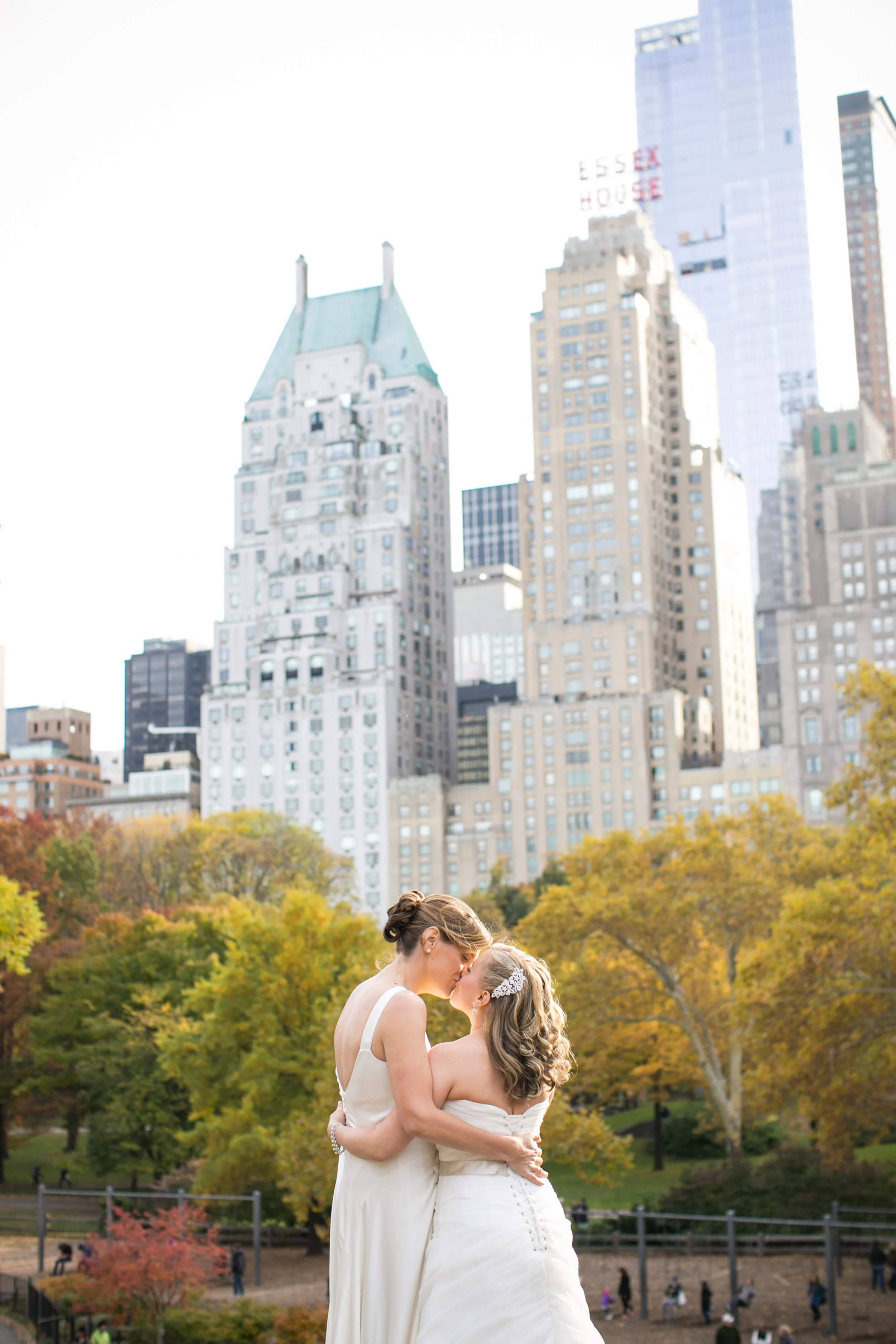 Two women in wedding dresses embrace and kiss in a park with colorful autumn trees, city skyscrapers in the background.