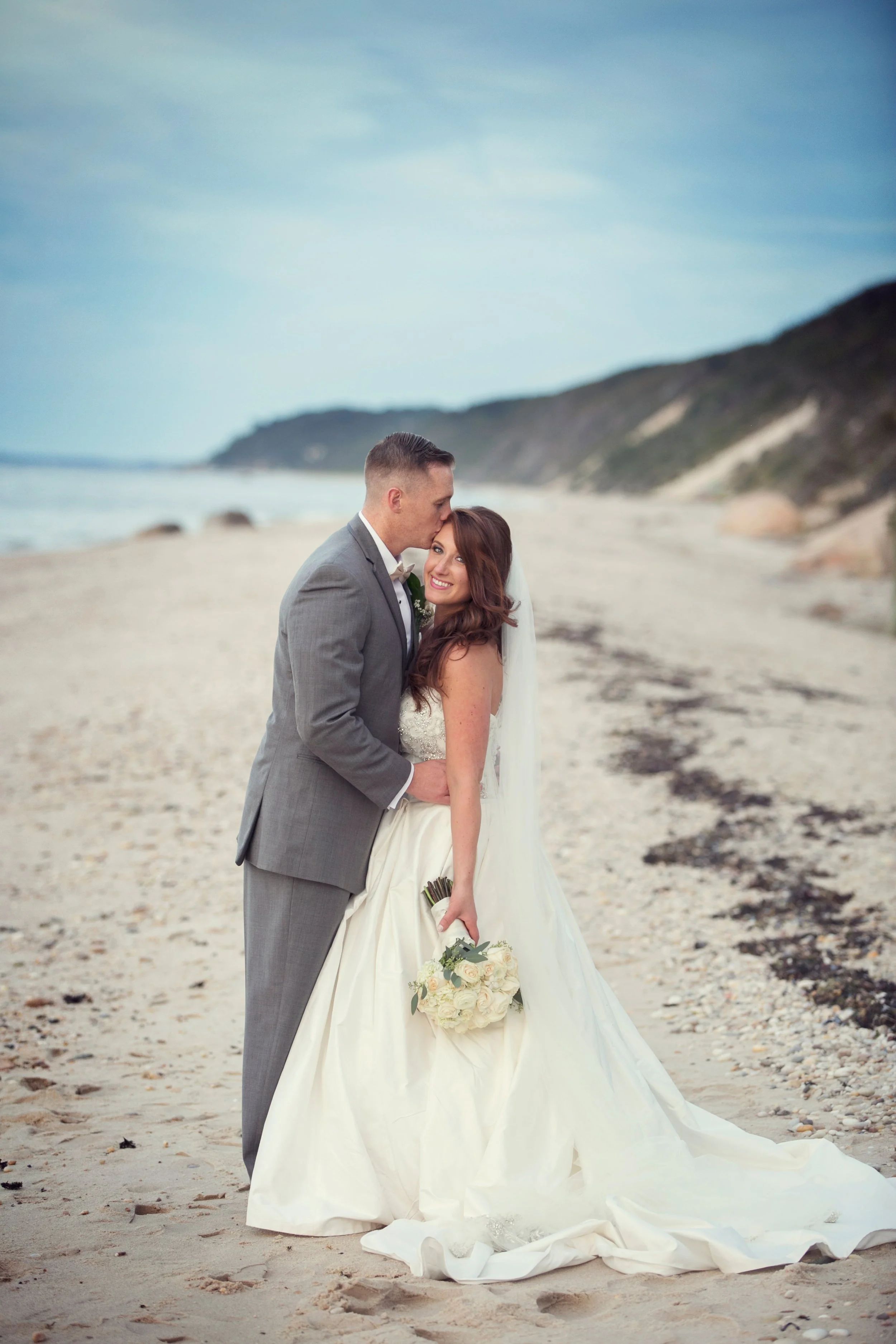 A bride and groom stand on a beach, with the groom kissing the bride's temple, and the bride smiling, holding a bouquet of white roses.