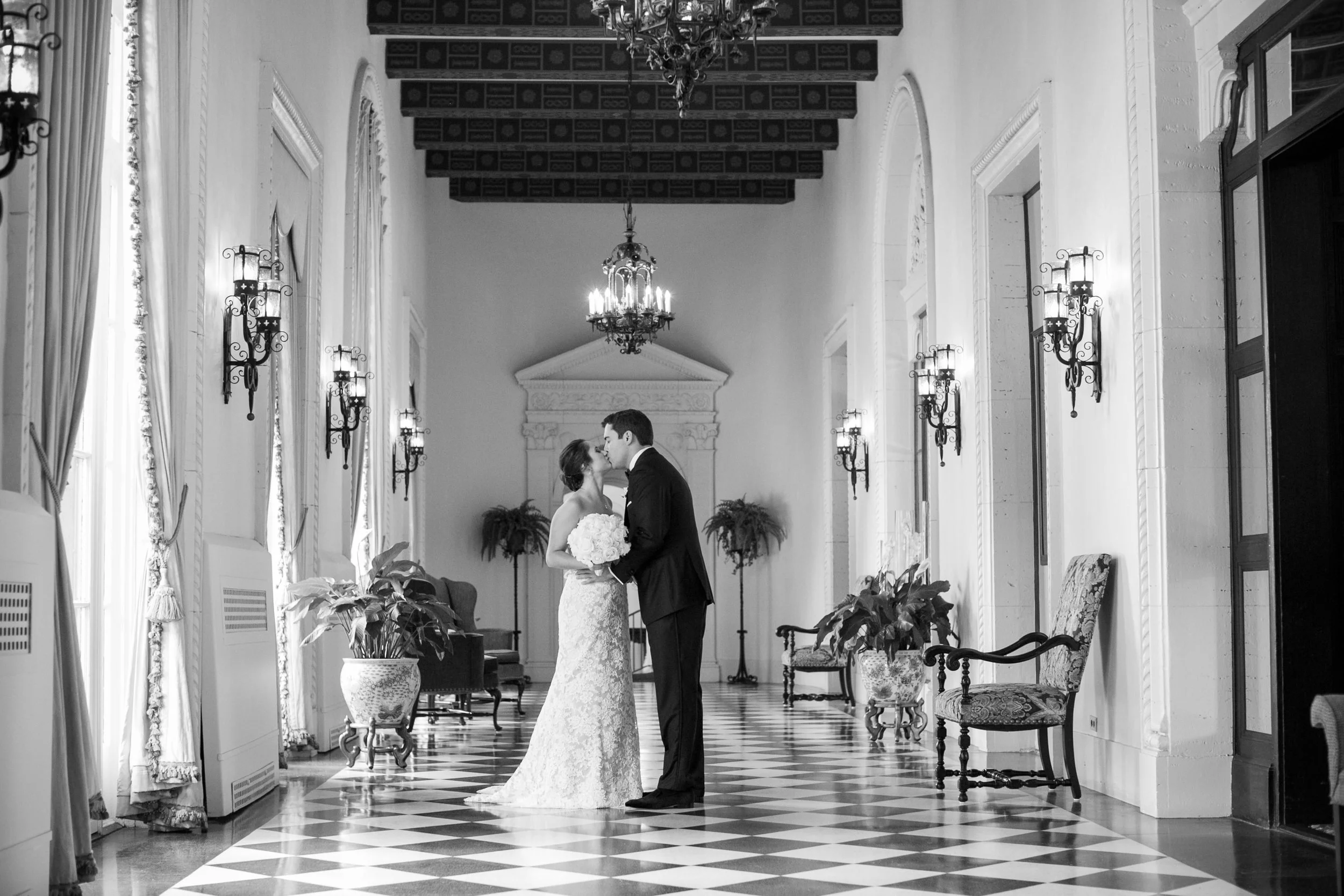 Black and white photo of a bride and groom standing in a grand hallway, sharing a kiss. The bride is holding a bouquet of flowers, and the groom is dressed in a suit. The hallway has tall windows with drapes, ornate wall sconces, a chandelier, and cl