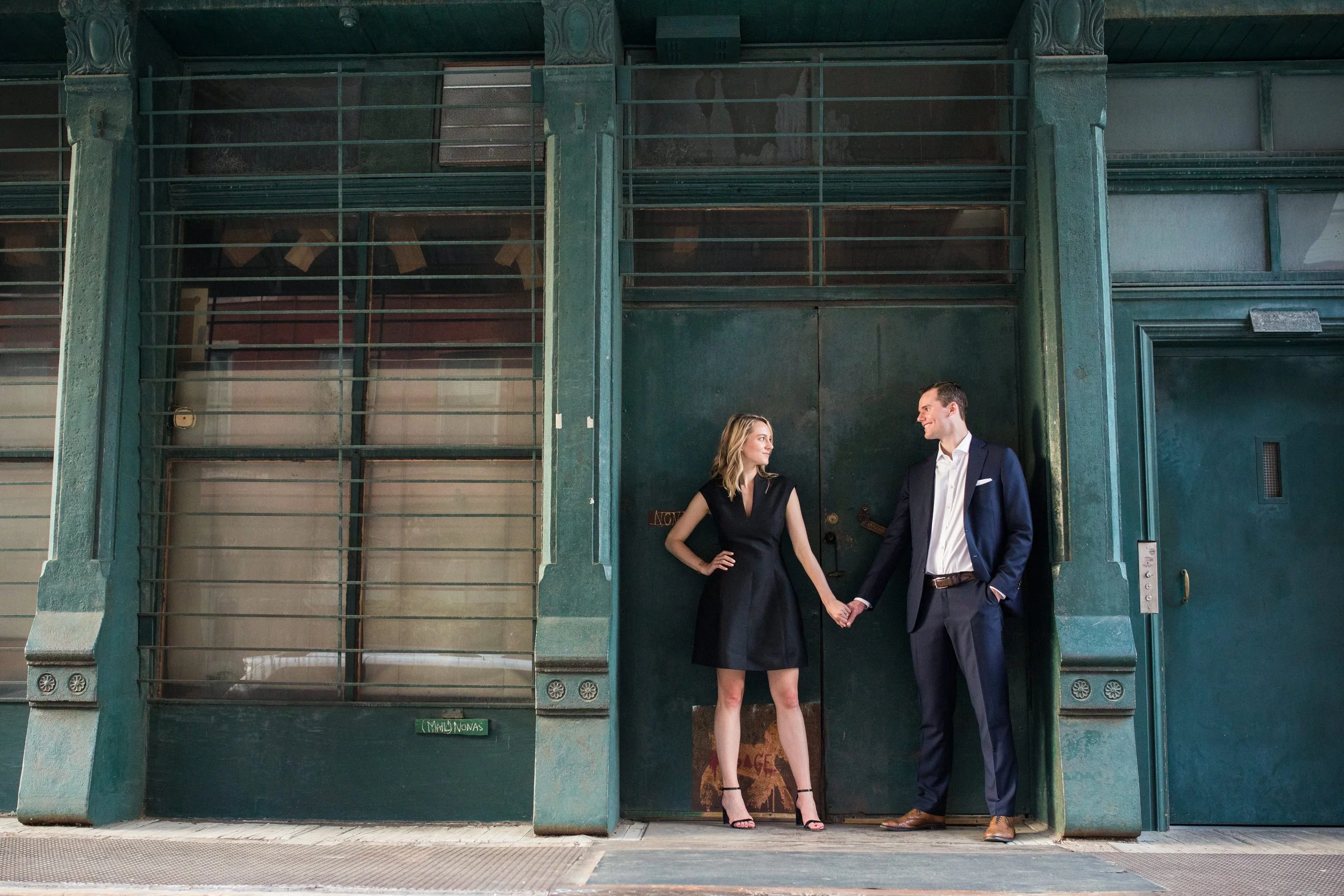 A man and woman holding hands and looking at each other while standing outside a green industrial door.