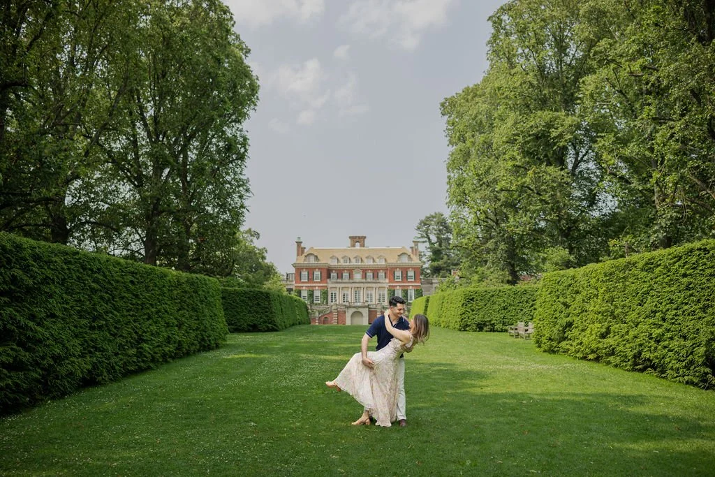 A couple dancing on a large, grassy lawn with a mansion and trees in the background.